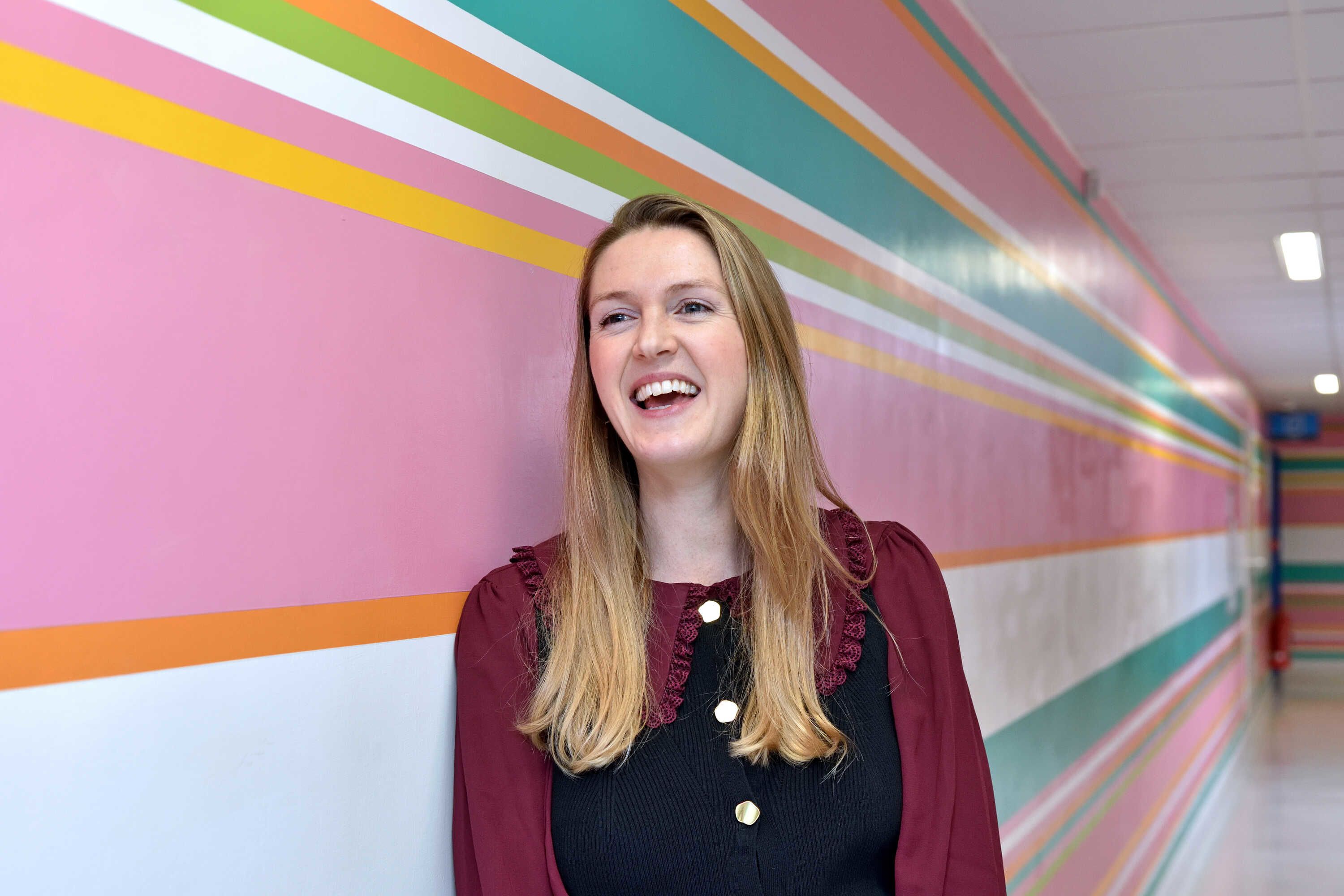 Anna smiling into the distance over the photographers left shoulder while leaning against a white, turquoise, pink and orange striped wall.