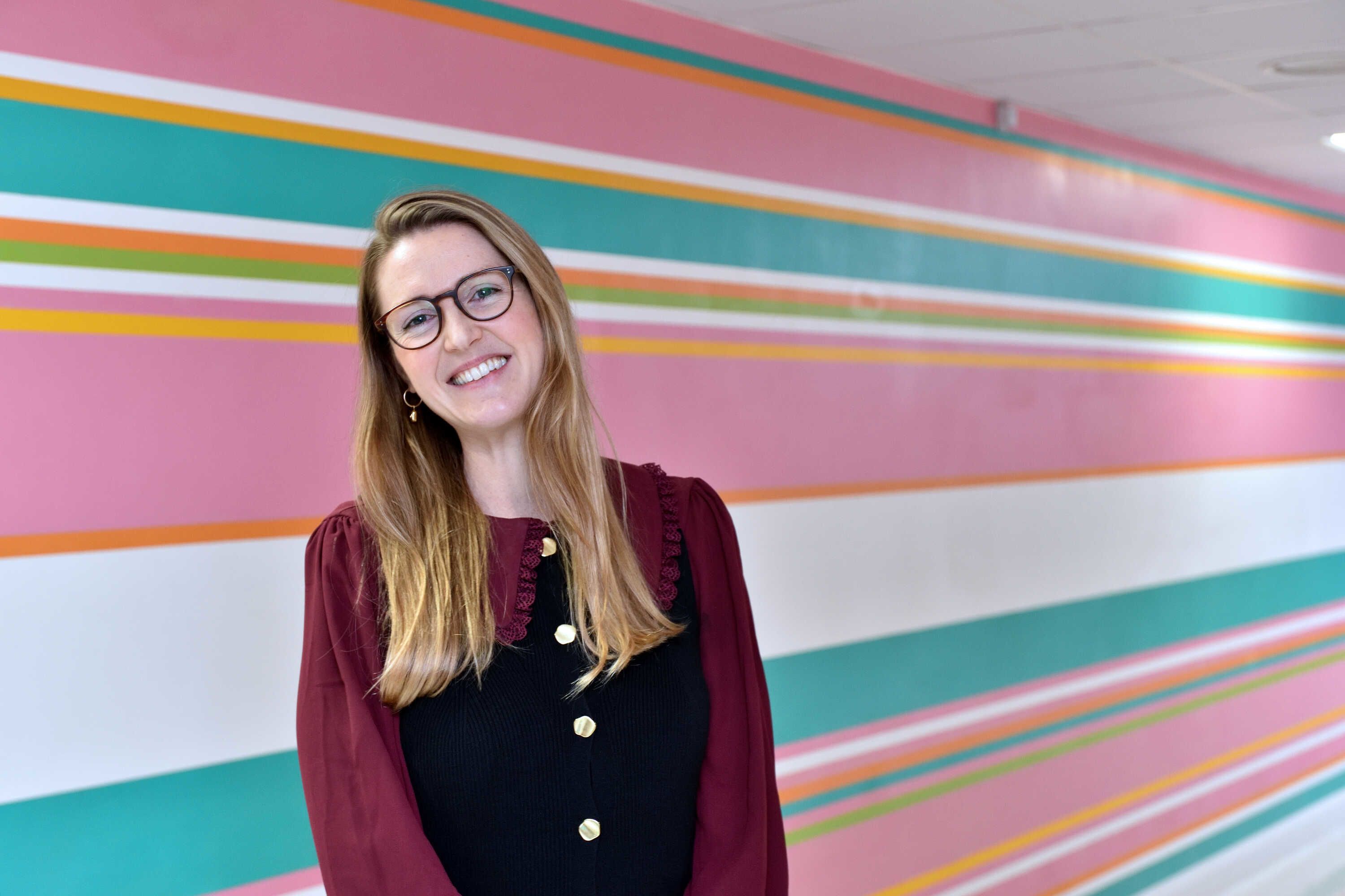 Anna wearing her glasses and smiling into the camera with a pastel pink, turquoise, white and orange striped wall in the background.