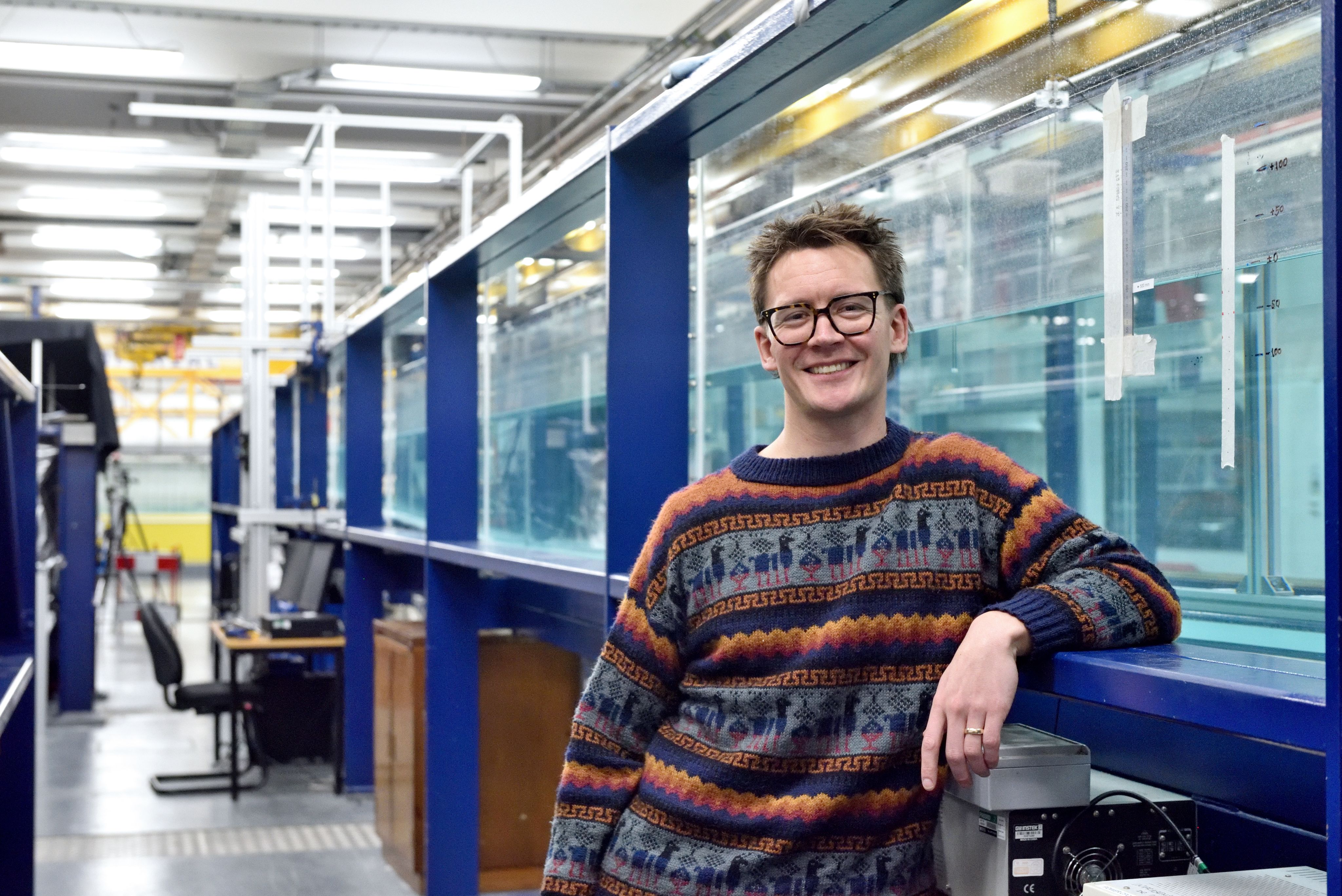 Ben with one elbow up leaning against a massive water tank and smiling at the camera, wearing a multicoloured jumper and glasses.