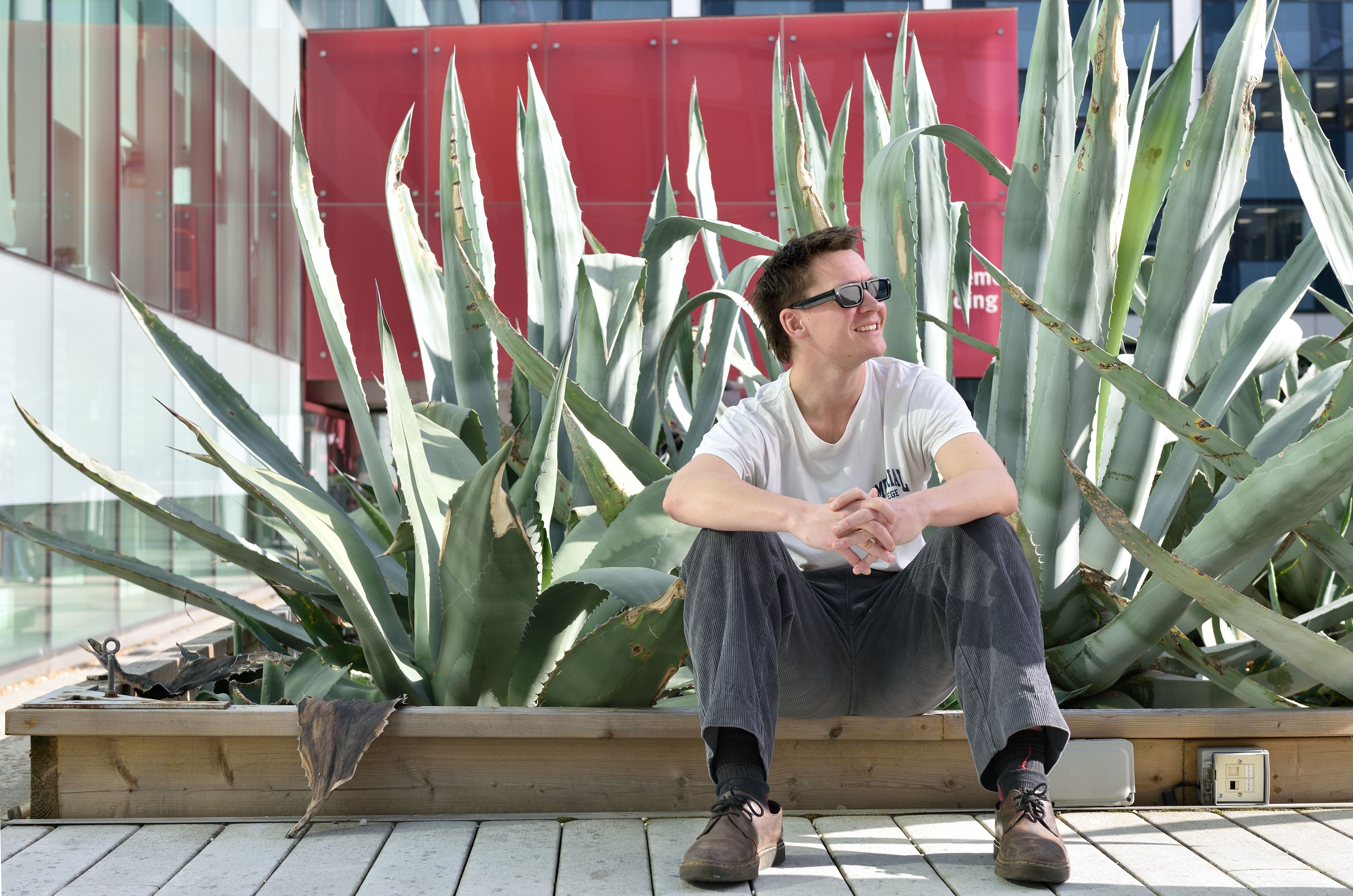 Ben sitting on a wooden garden wall in front of a large cactus, wearing his sunglasses and staring to the right of the photographer.