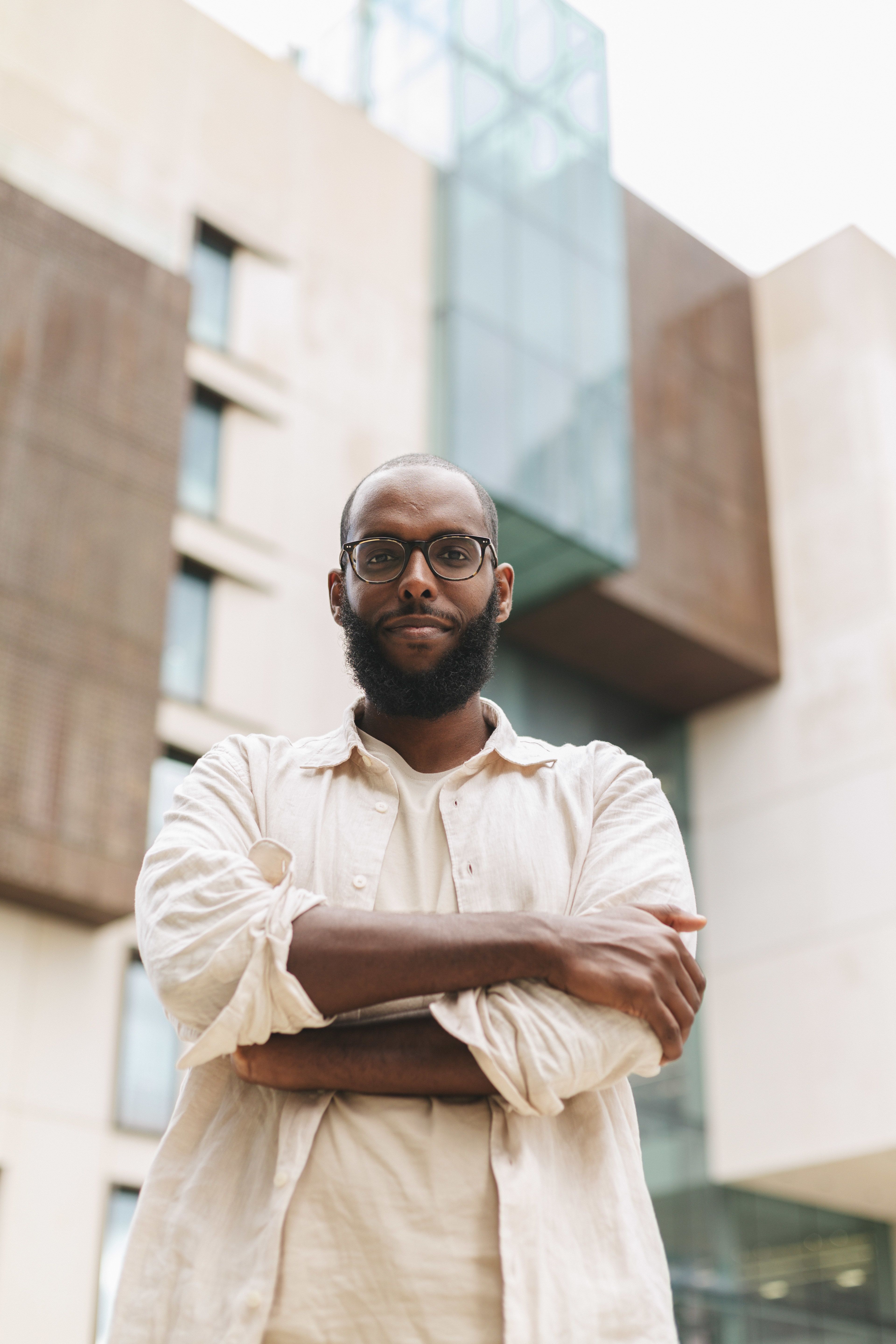 Faysal standing in front of the building at Imperial College London his PhD was based in, with his arms crossed, looking directly at the camera.