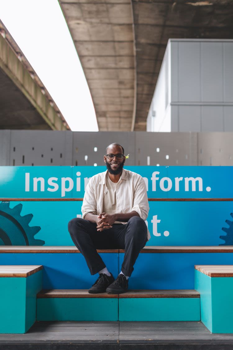 Faysal sat on a bench, against a blue background, smiling and looking at the camera.