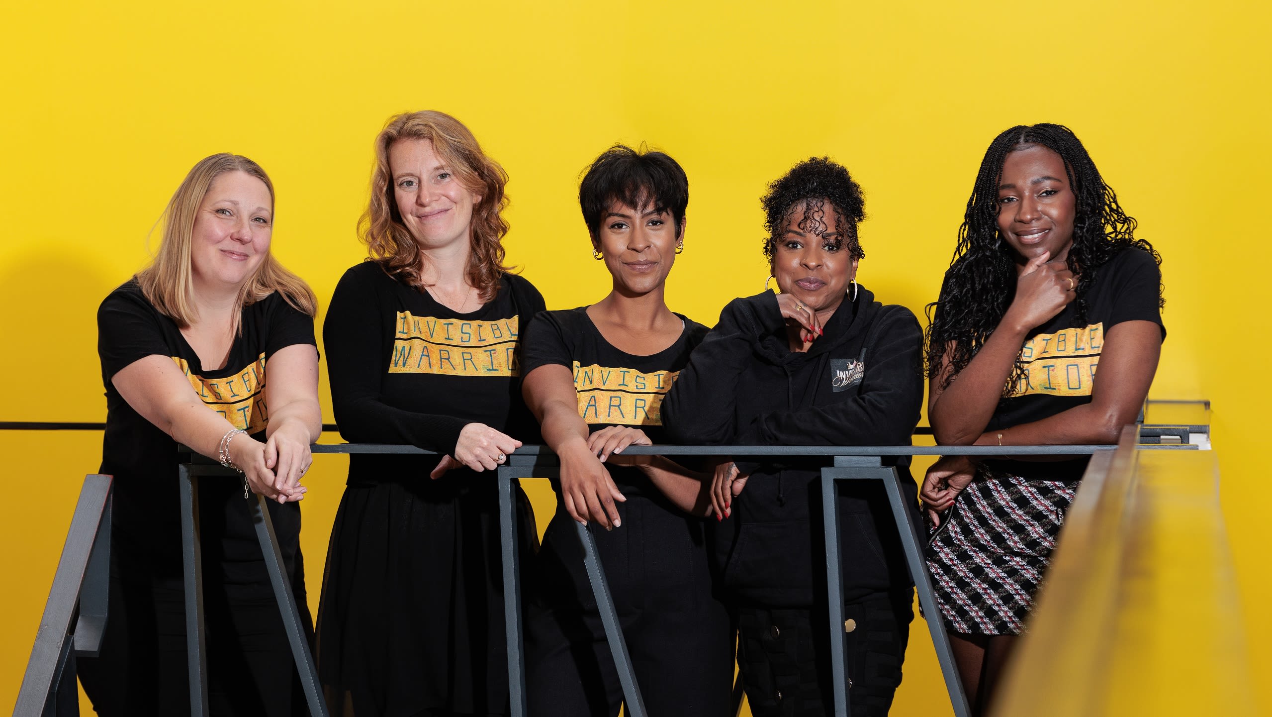 The Invisible Warrior team, five women standing against a yellow background, smiling