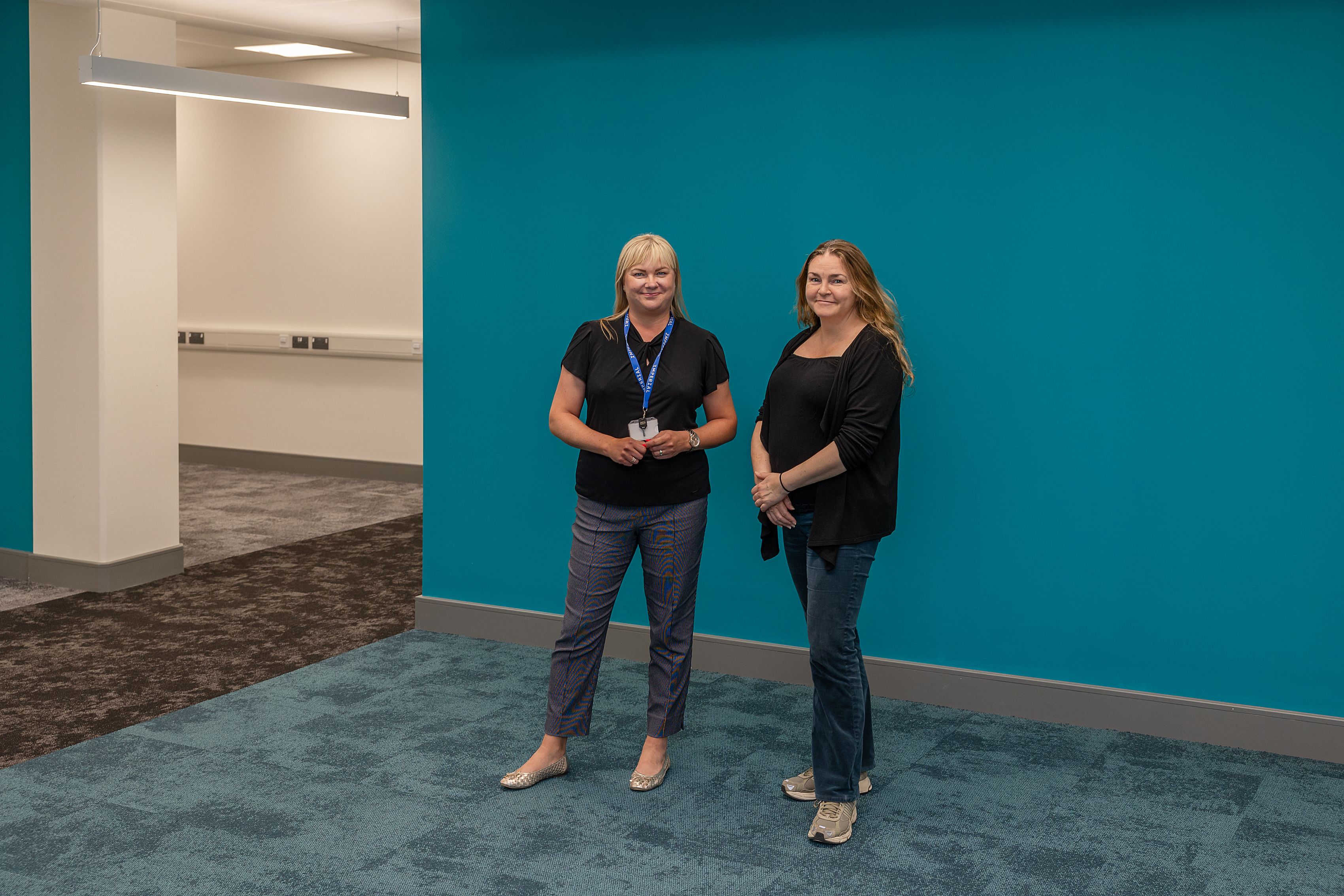 Project delivery Project Manager Monika Kojak and Faculty project Manager Same Tolhurst standing together in fron ot a teal coloured wall in the empty communal office space