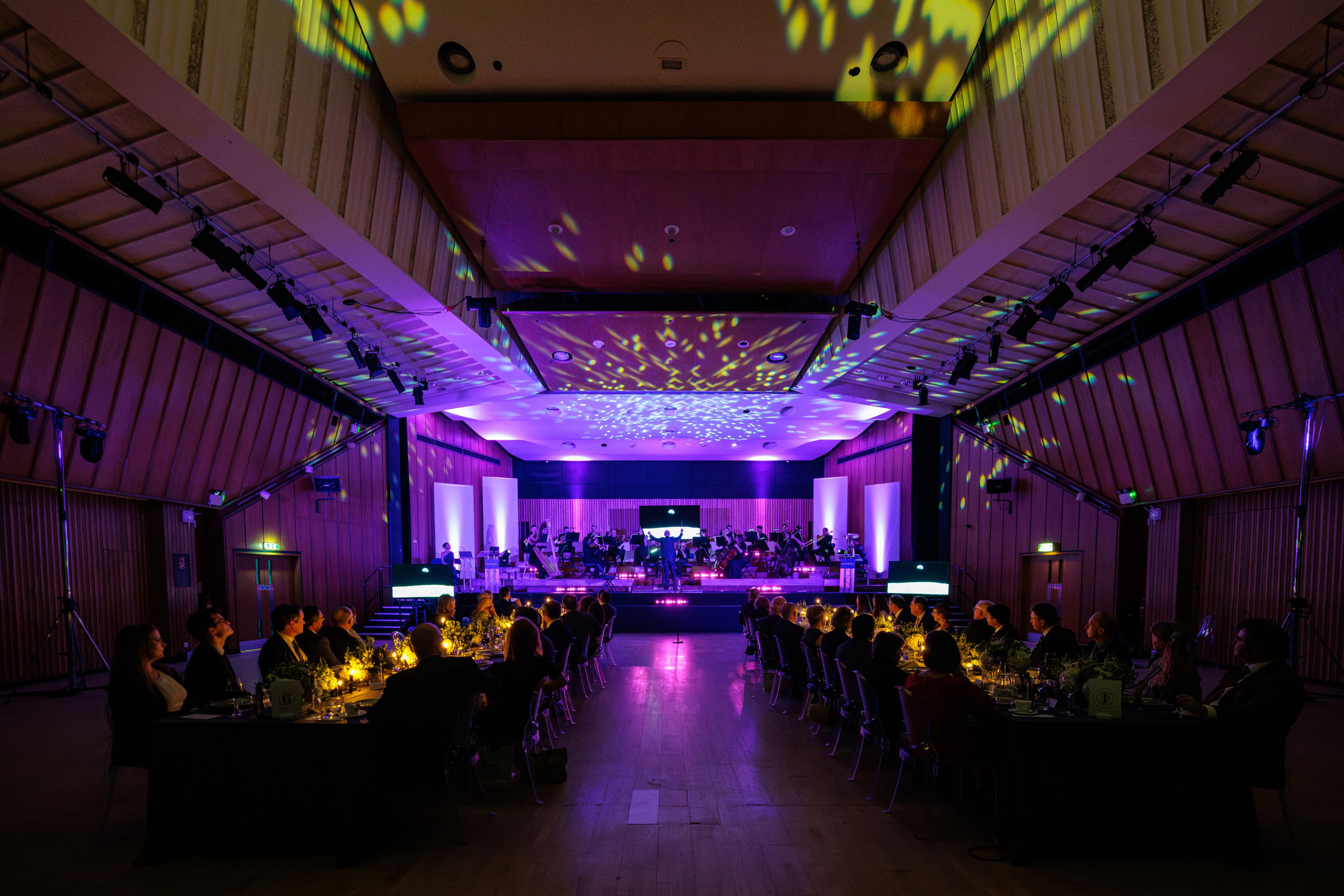 A photograph of the Great Hall set up for the Circle of Benefactors dinner in 2024. The room is dark and illuminated with purple and yellow lights.