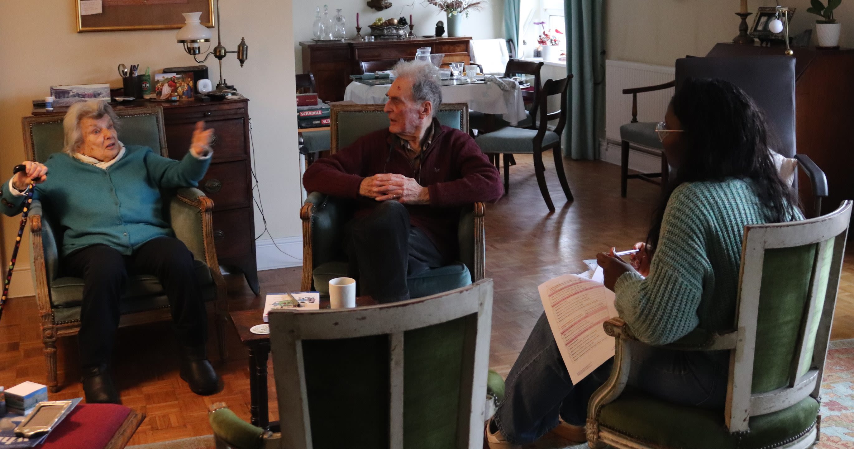 A picture of two elederly dementia patients in their living room being visited by a researcher for an assessment