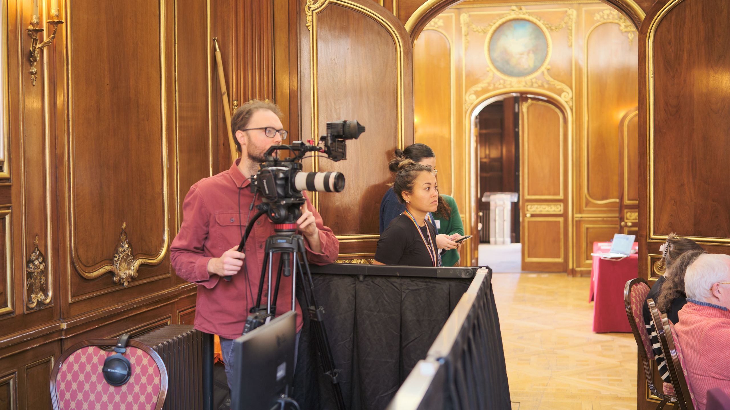 a person filming in an ornate room while a woman looks on