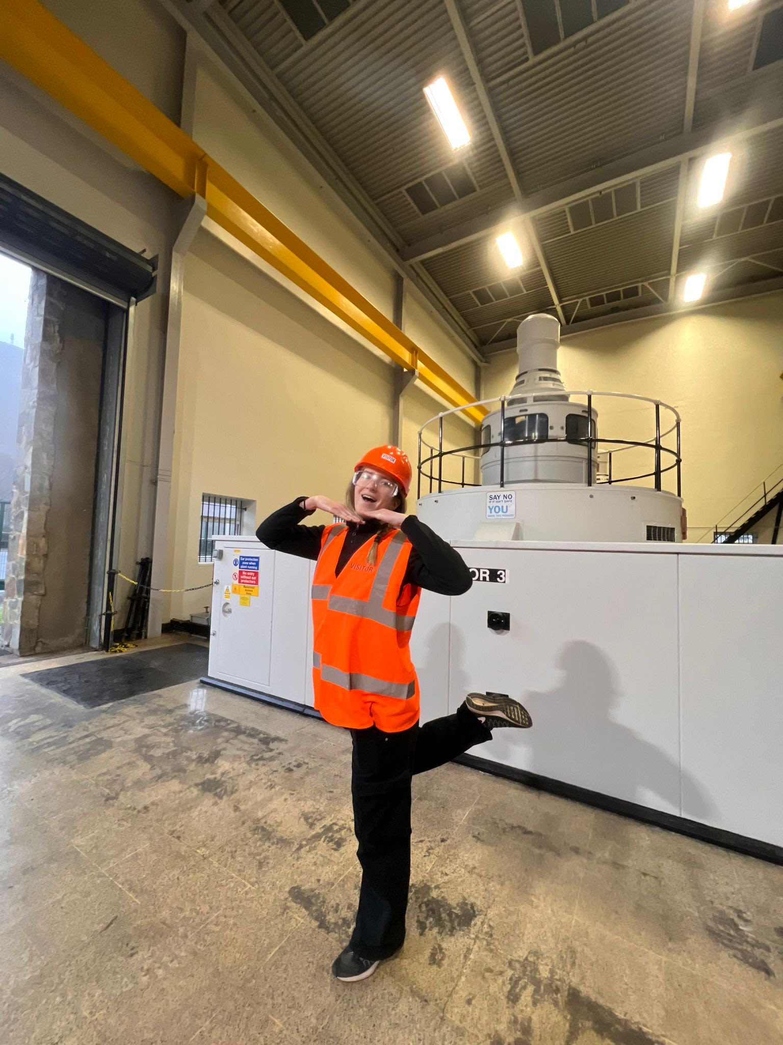 Alexandra wearing an orange hard hat and reflective vest posing in front of large machinery