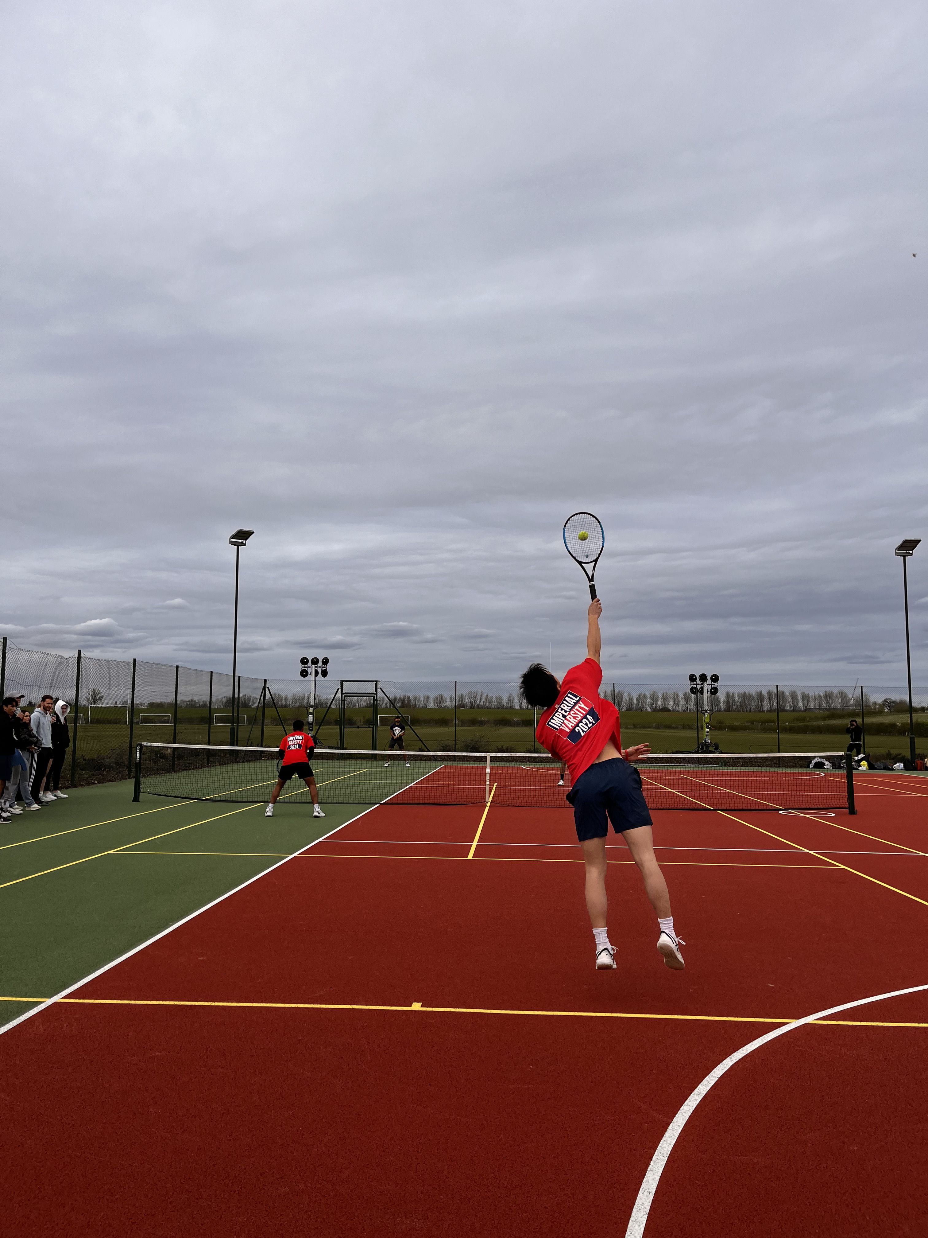 Action shot of Andrew reaching up in the air to hit a tennis ball on a tennis court