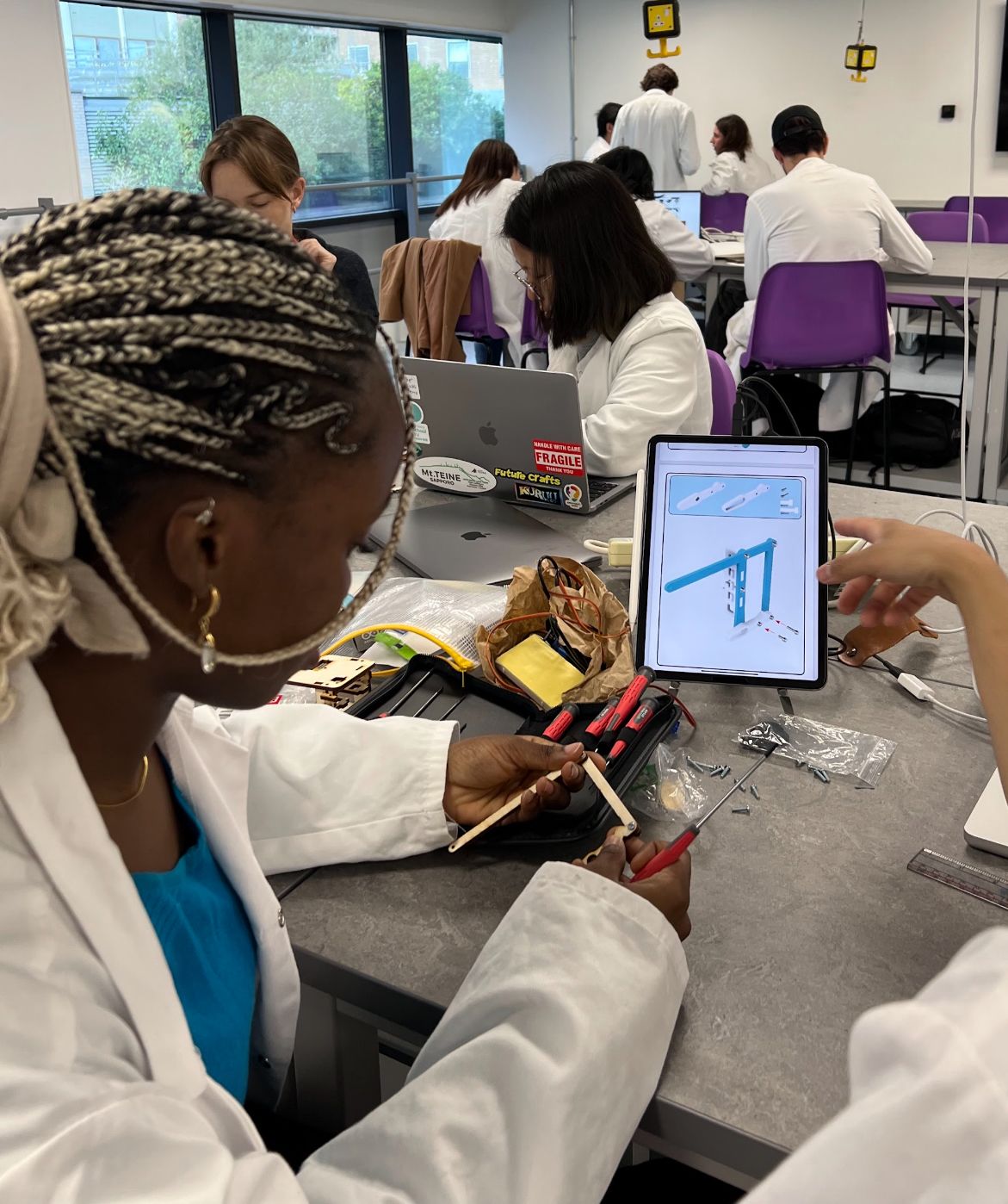 Ifeoluwa wearing a lab coat and working on a design project in a classroom with other Innovation Design Engineering students in the background
