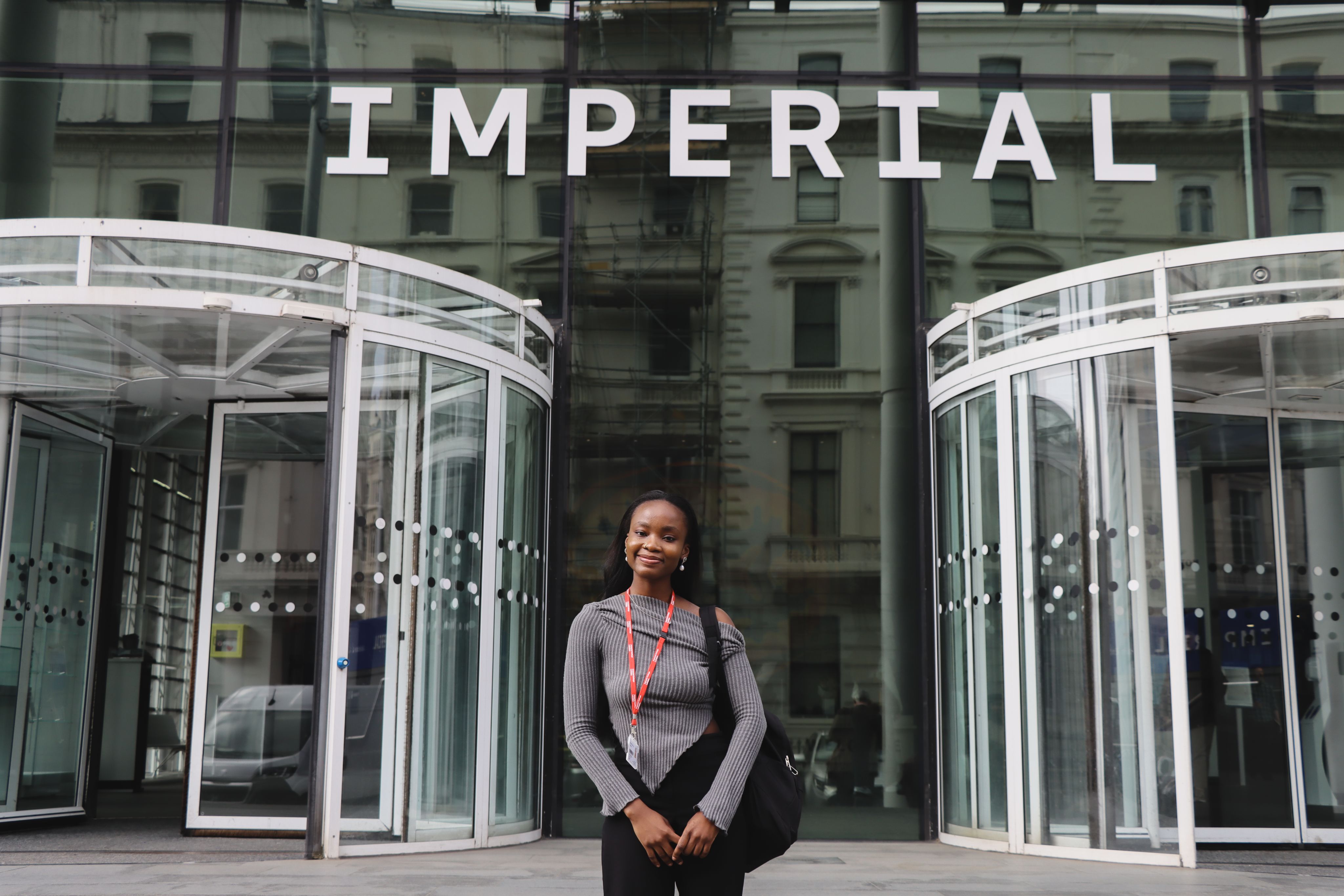 Ifeoluwa standing in front of the Imperial logo in between the revolving doors at the main entrance of Imperial's South Kensington Campus
