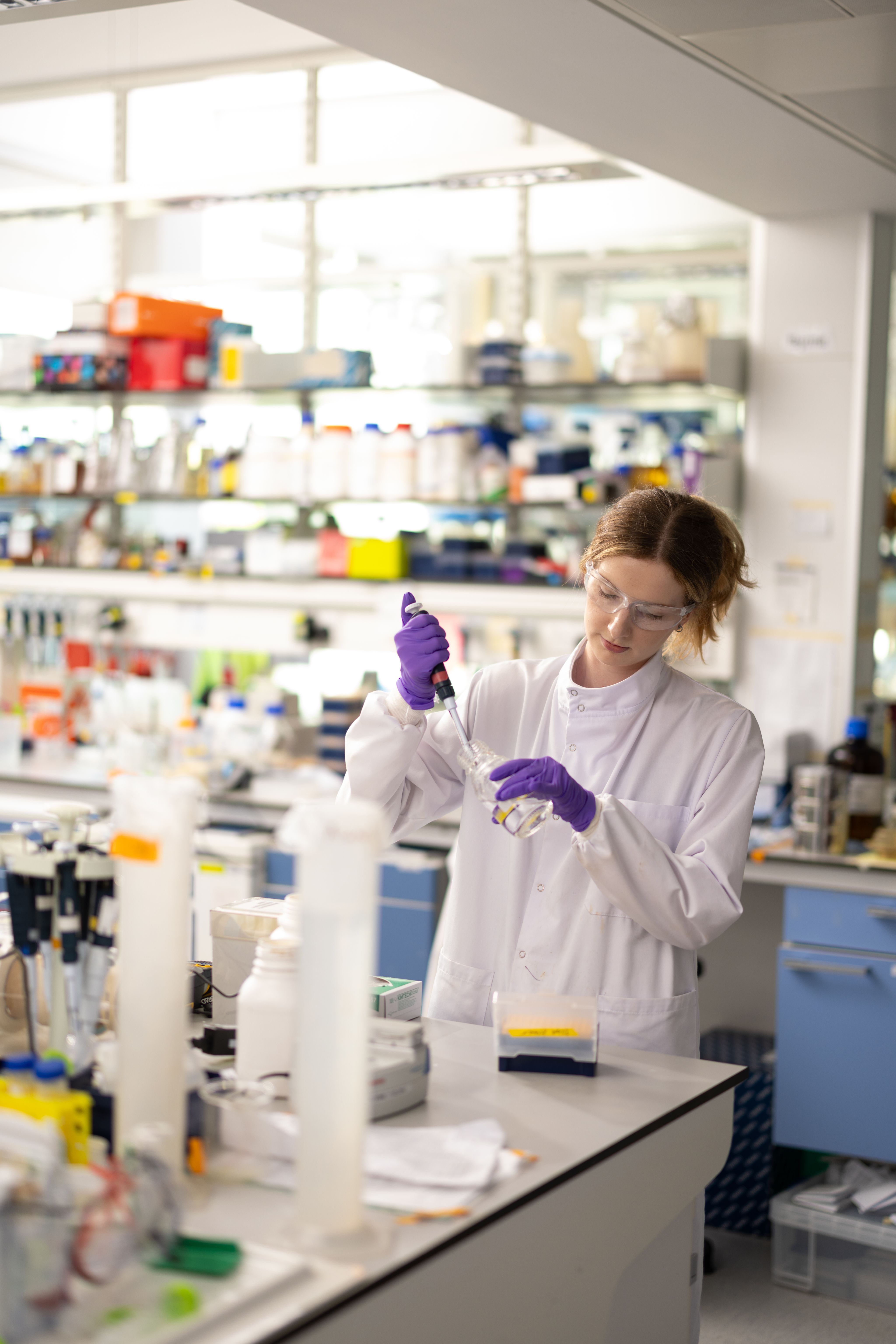 Katie in a lab using a pipette, wearing a white lab coat, purple gloves and safety goggles