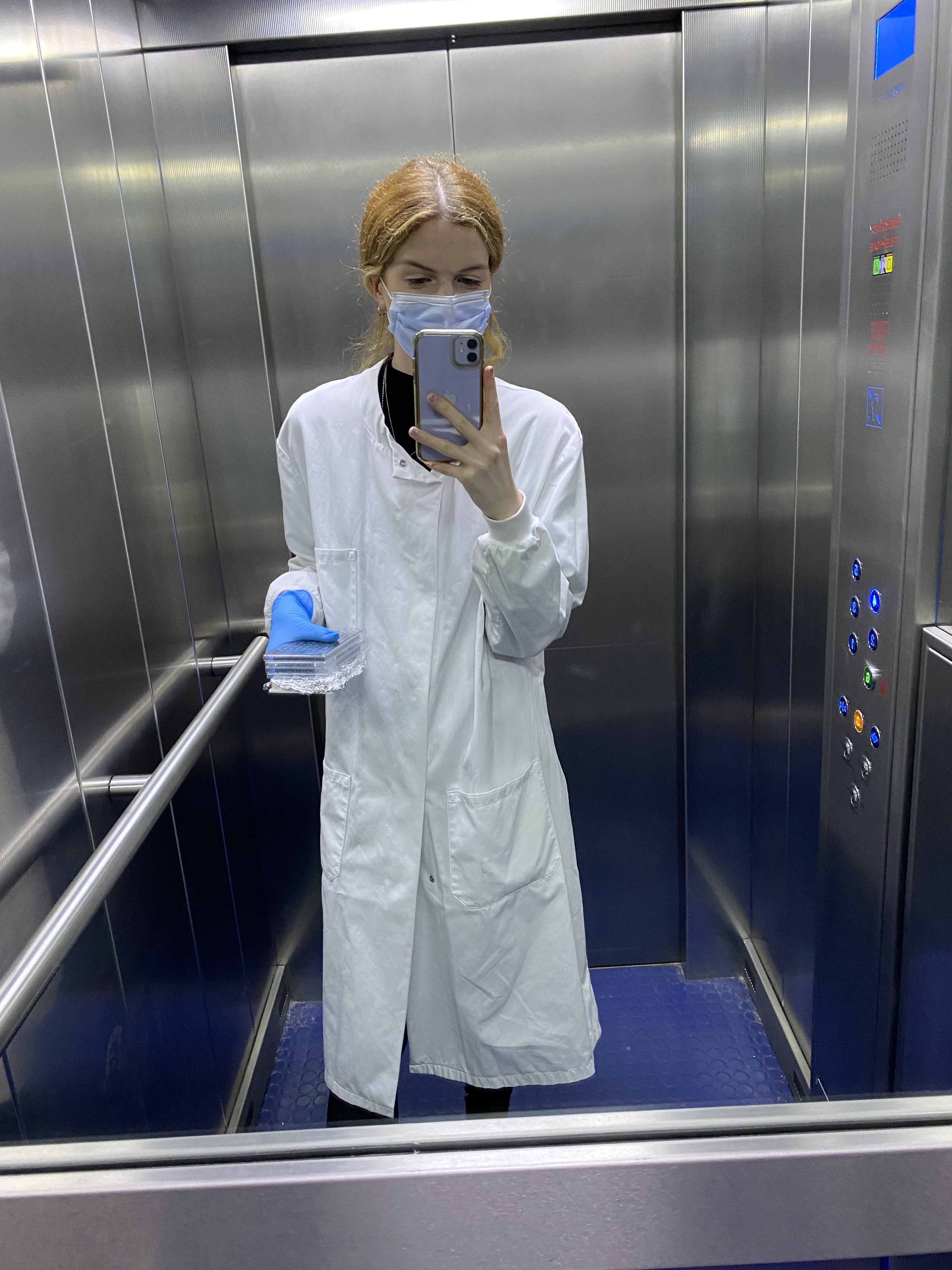 Mirror selfie of Katie in a lift, wearing a lab coat, a face mask and gloves