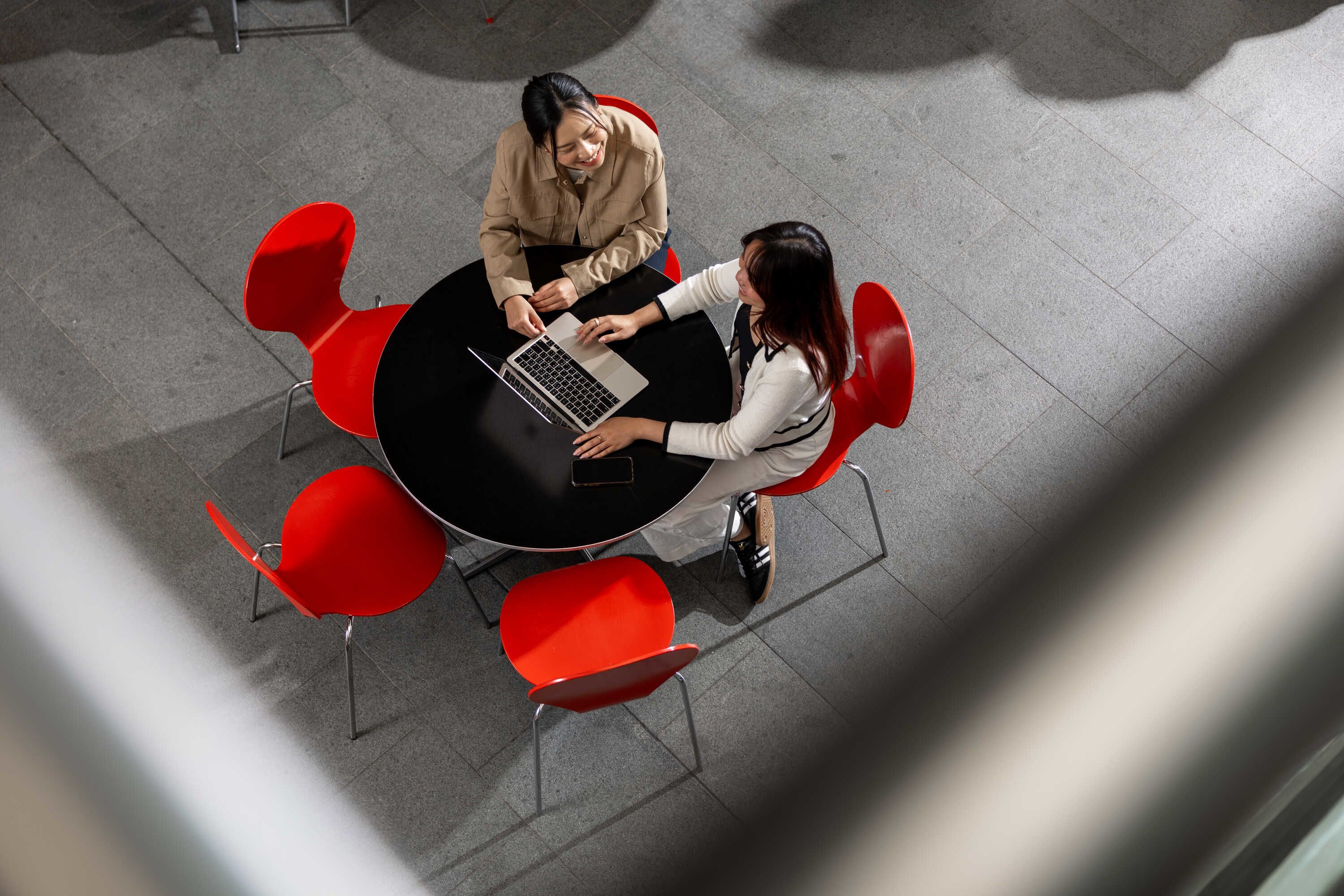 Bird's eye view of Rose sitting at a round table with red seats with a friend looking at an open laptop