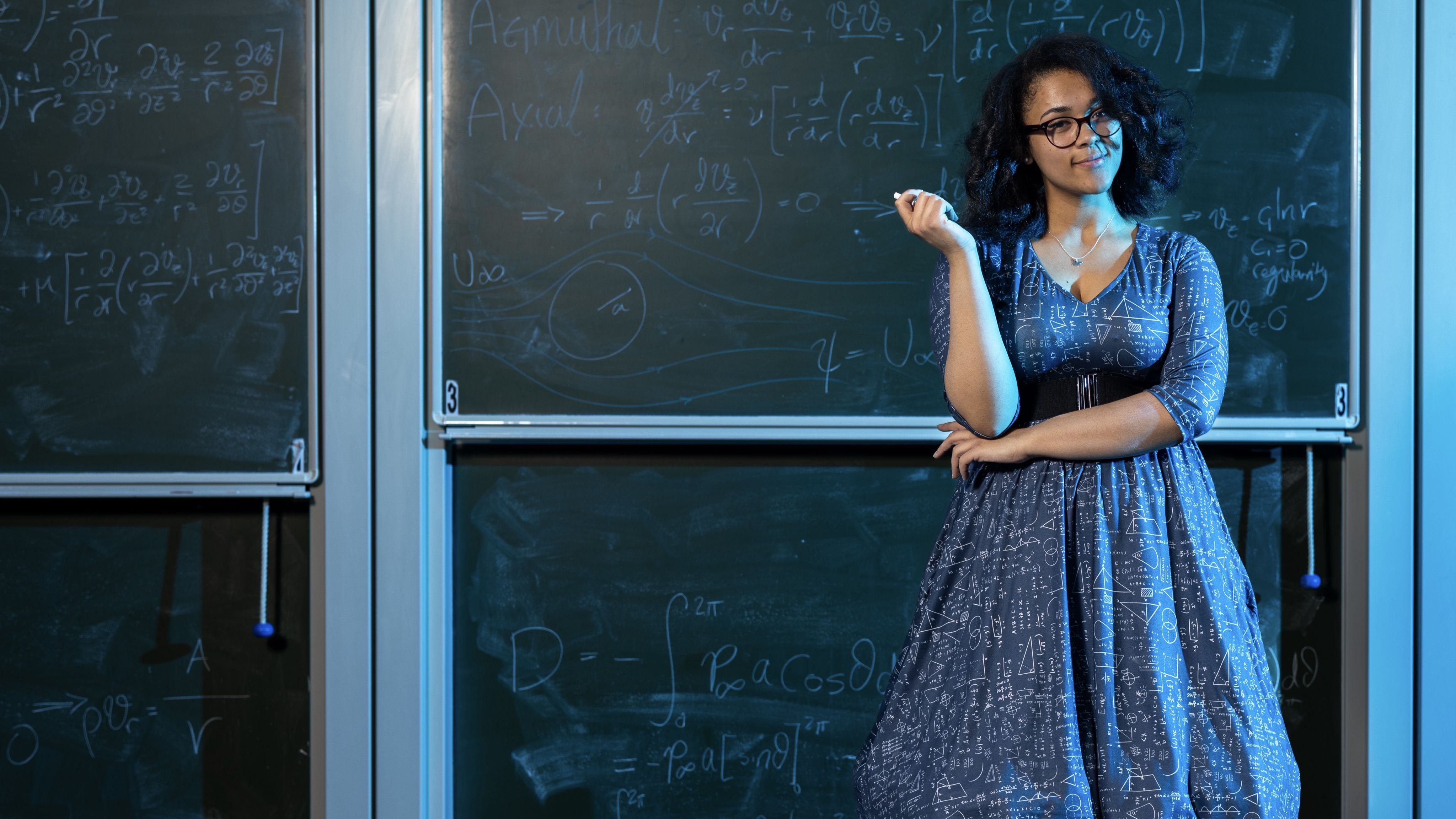 Vanessa holding some chalk and standing in front of a blackboard with maths equations on it
