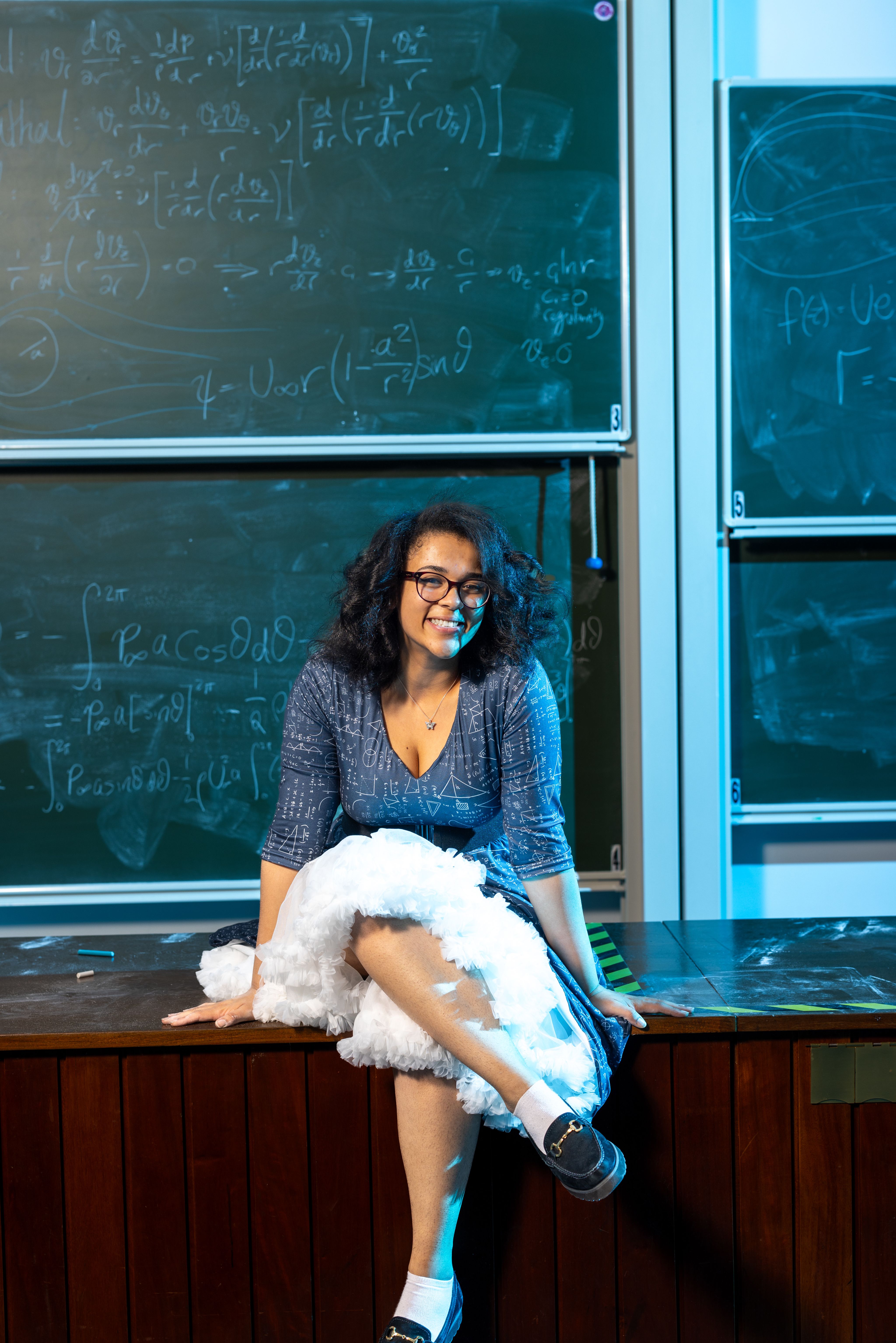Vanessa sitting on a desk in front of a blackboard with equations on it