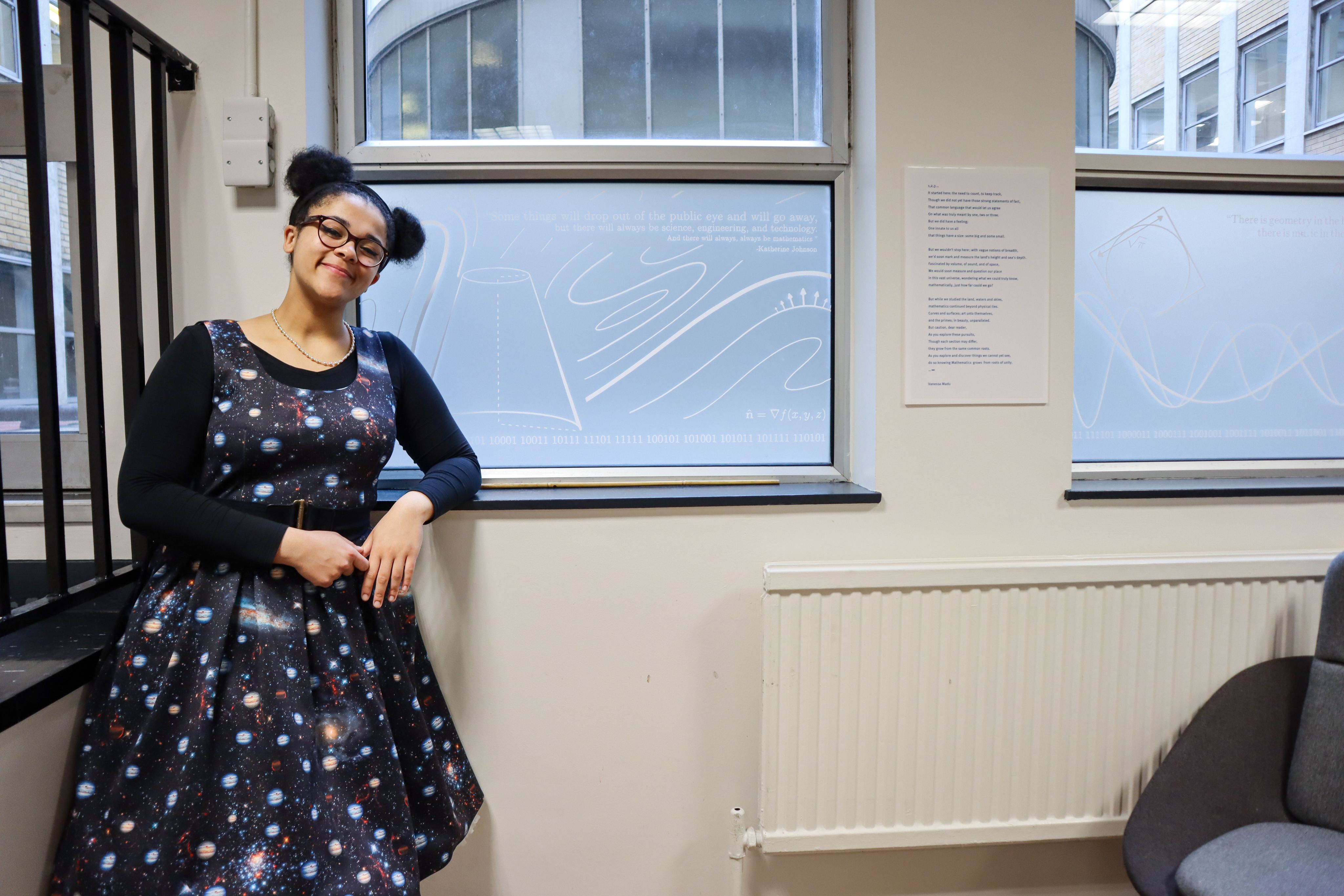 Vanessa standing in front of a window featuring her mathematical designs in Imperial's Huxley Building