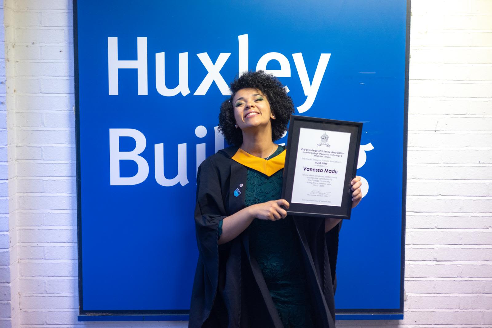 Vanessa in a graduation gown holding a certificate in front of a sign saying 'Huxley Building'