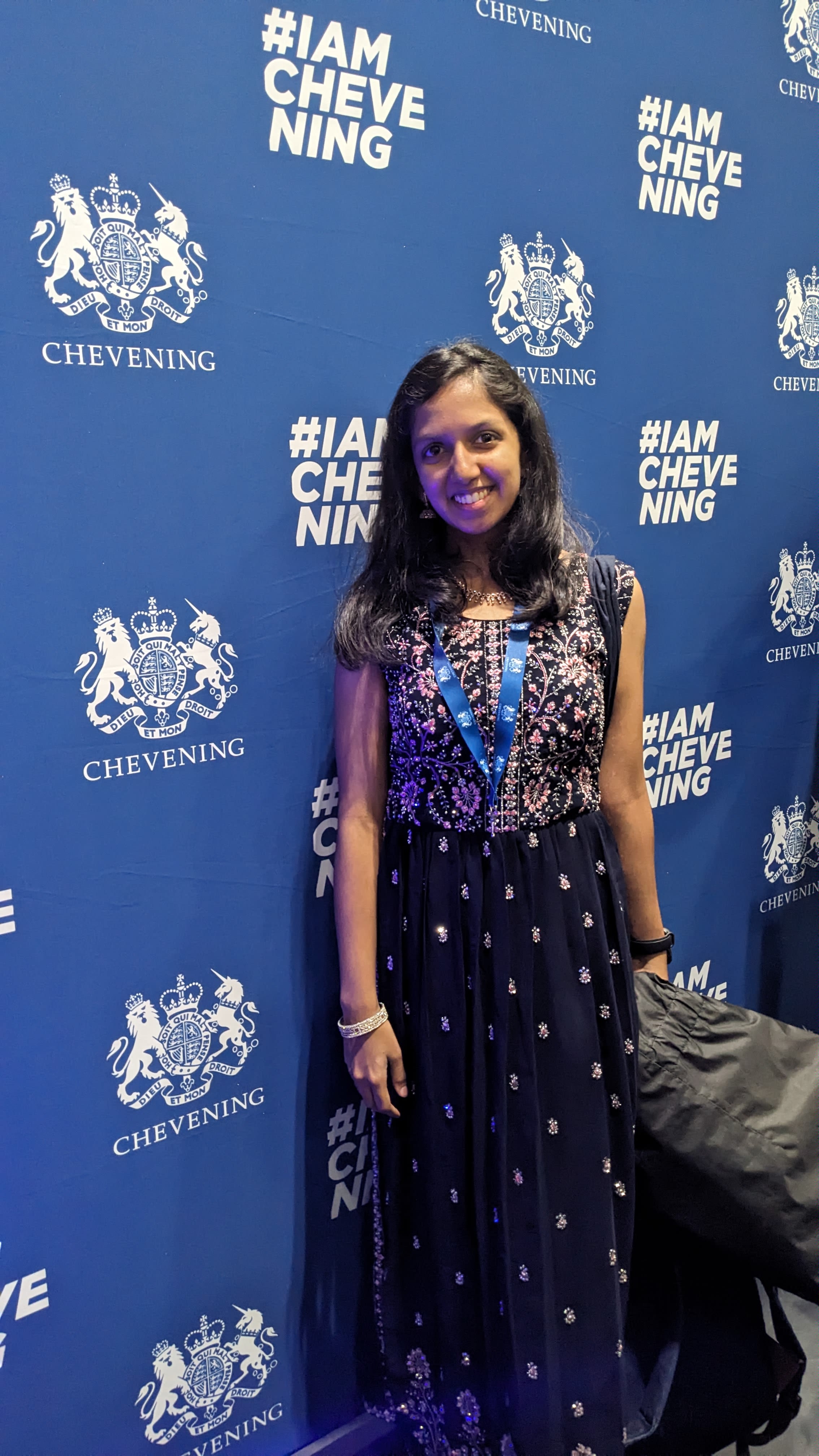 Vanya standing in front of a photograph wall with the Chevening logo and text saying 'I Am Chevening'.