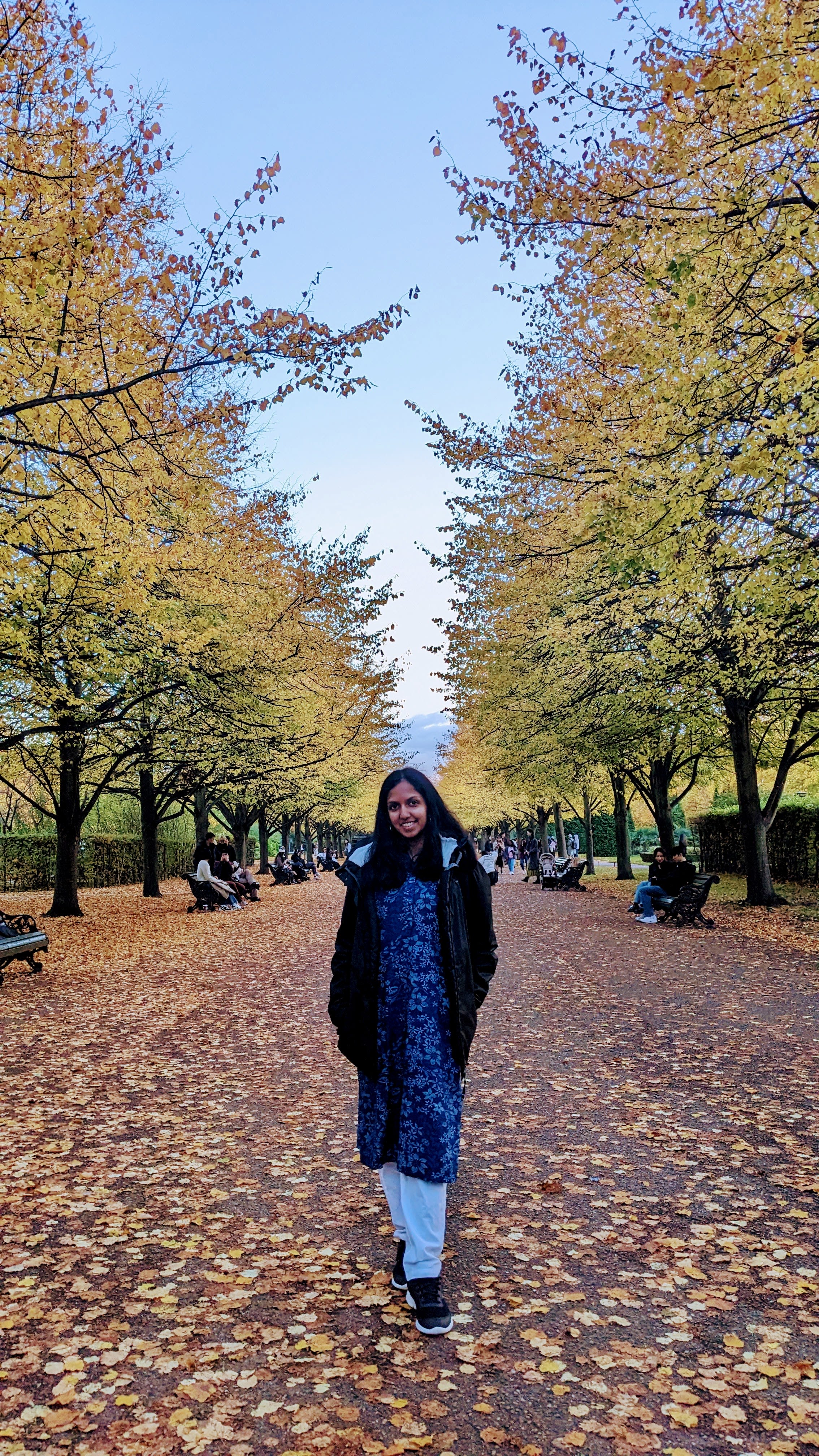 Vanya standing in the middle of a park with autumnal trees on either side of her