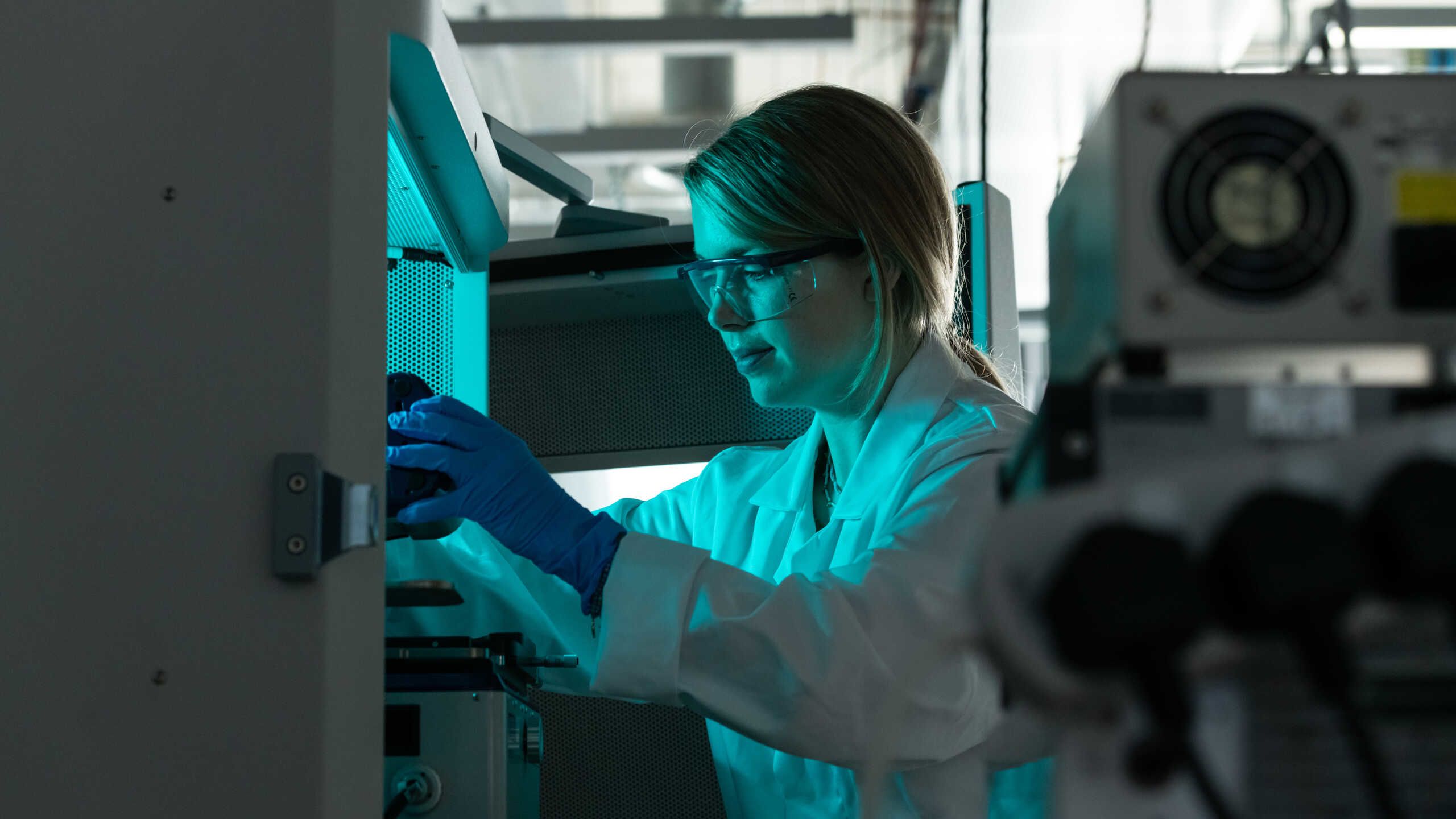 A researcher in a white coat and safety glasses is bathed in the blue light from a scientific instrument.