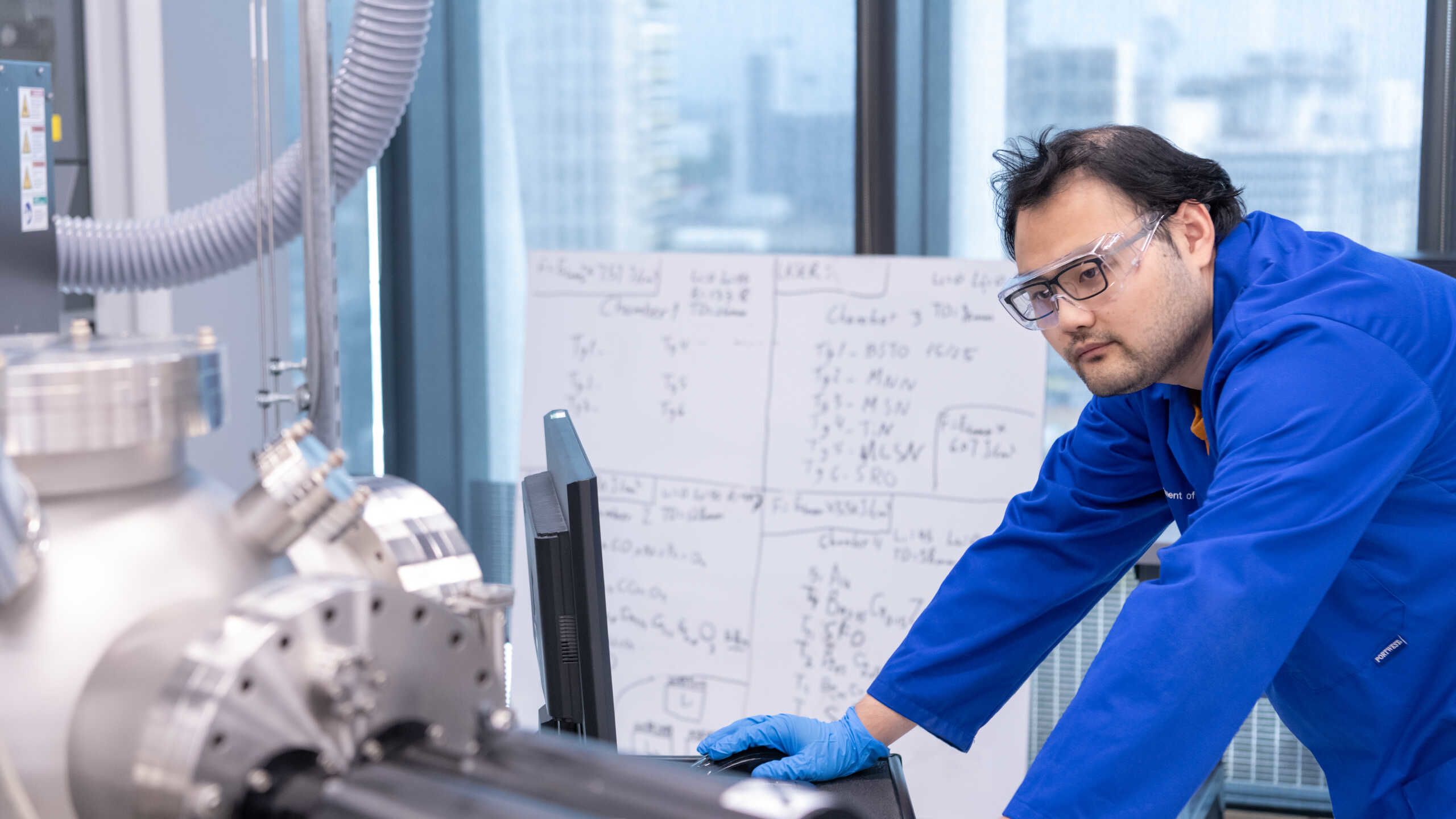 A technician in a blue coat looks at a screen. A white board covered in figures in the background.
