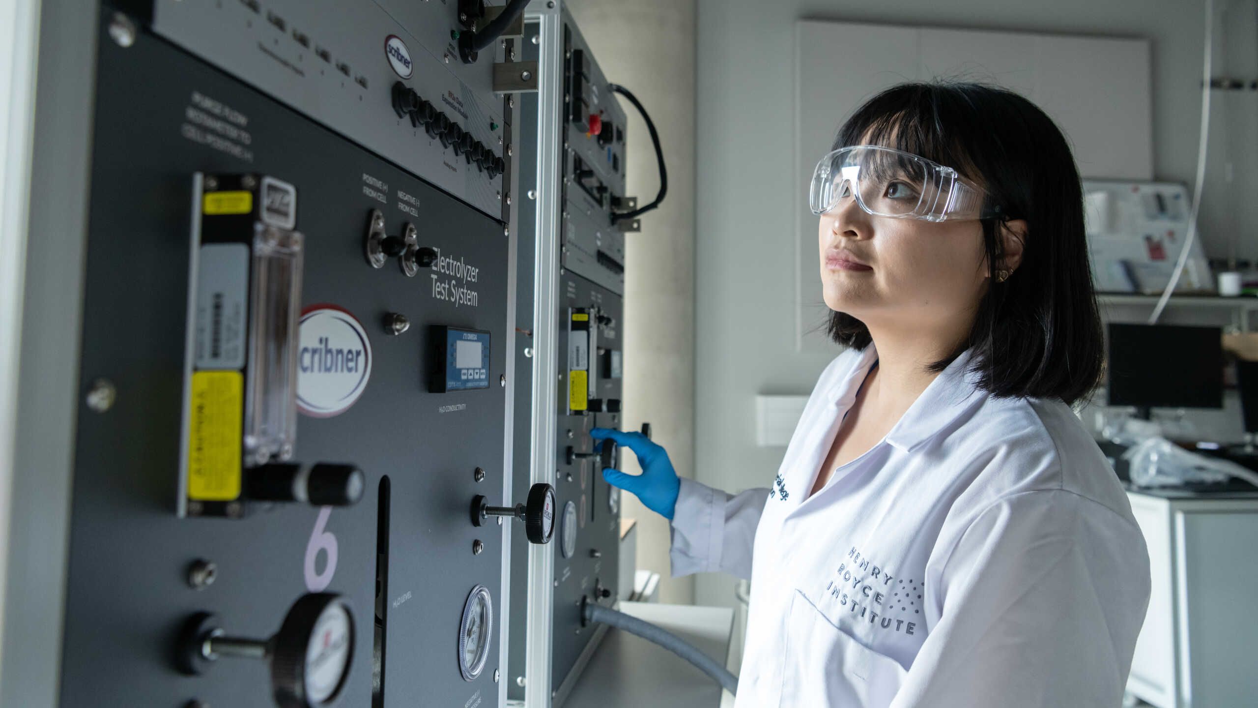 A researcher in white coat and safety glasses looks at an instrument.