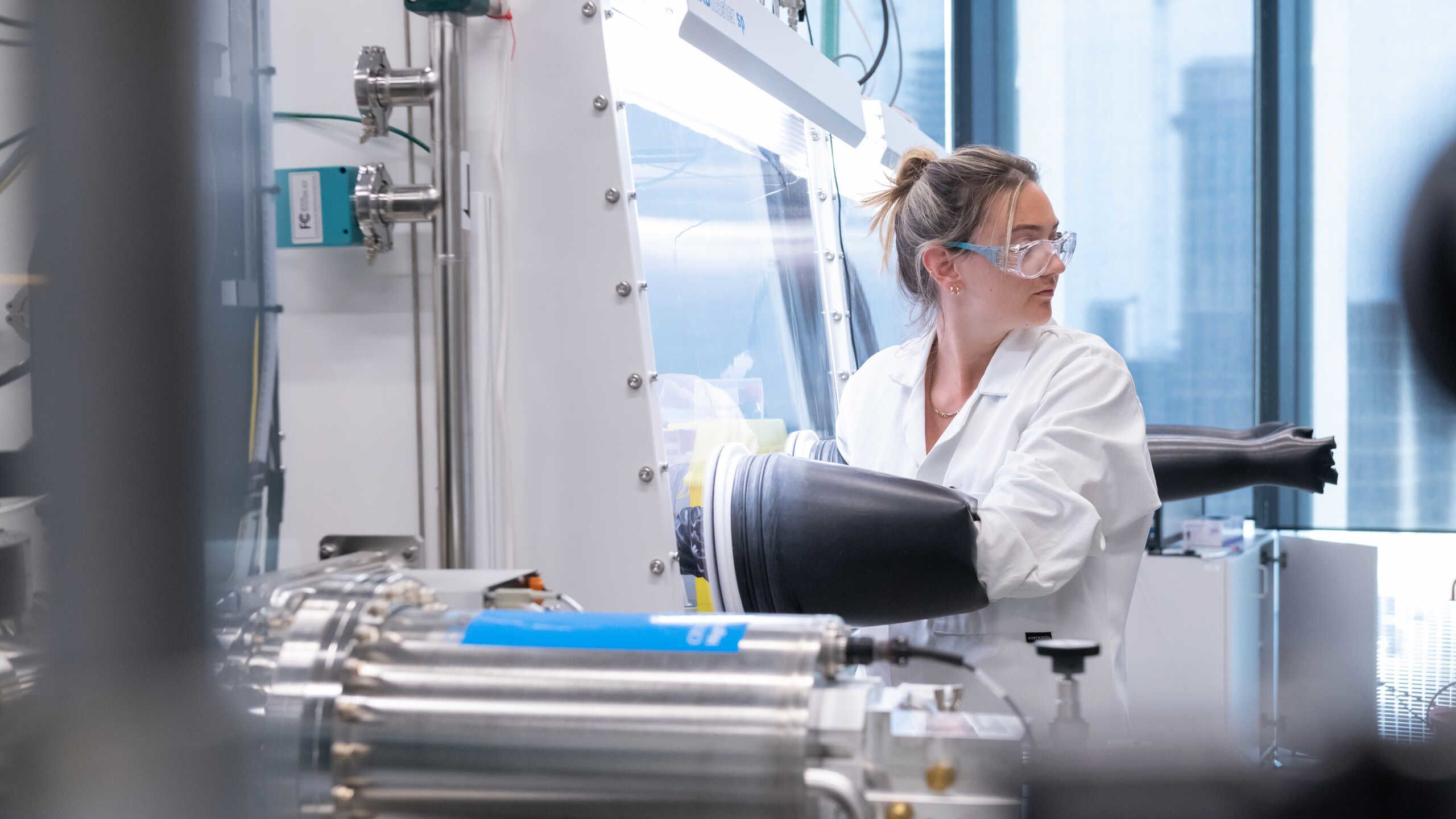 A reseaarchers in a white coat and safety glasses works at a glove box.