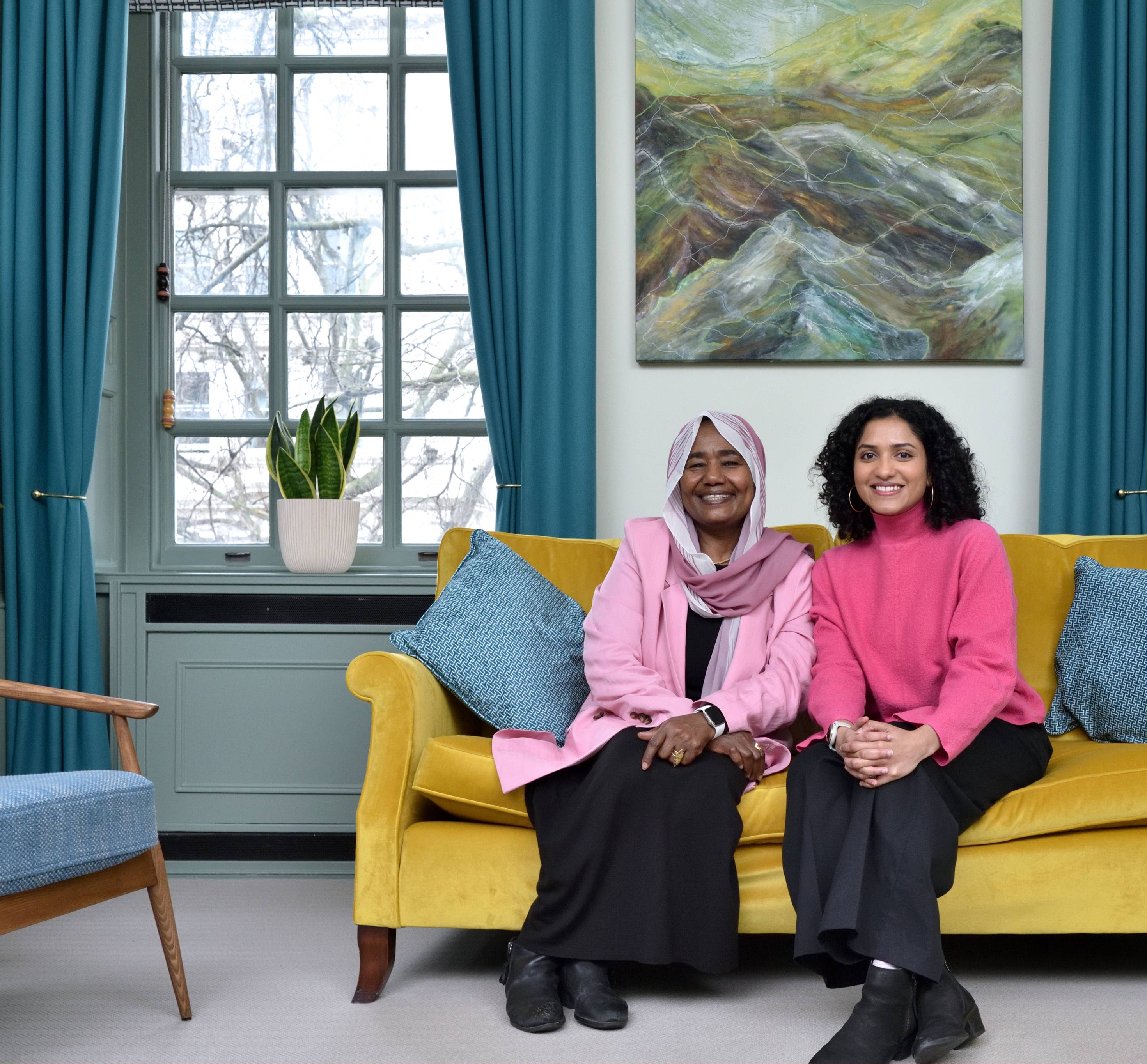 Sarindi Aryasinghe and Phayza Fudlalla sitting on a dark-yellow couch together facing the camera