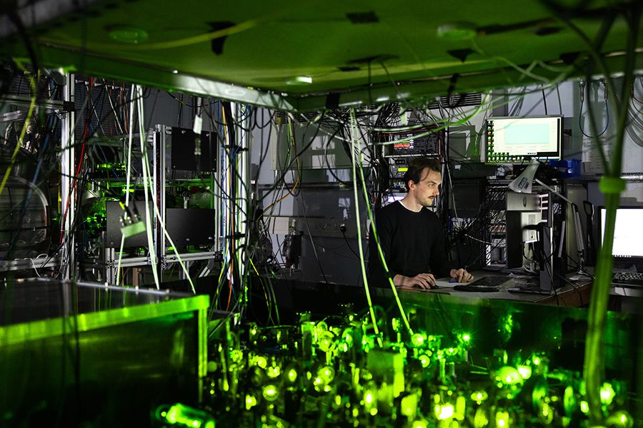 A scientist sits at a computer. The eEDM expeirment is in the foreground, lit by green laser light