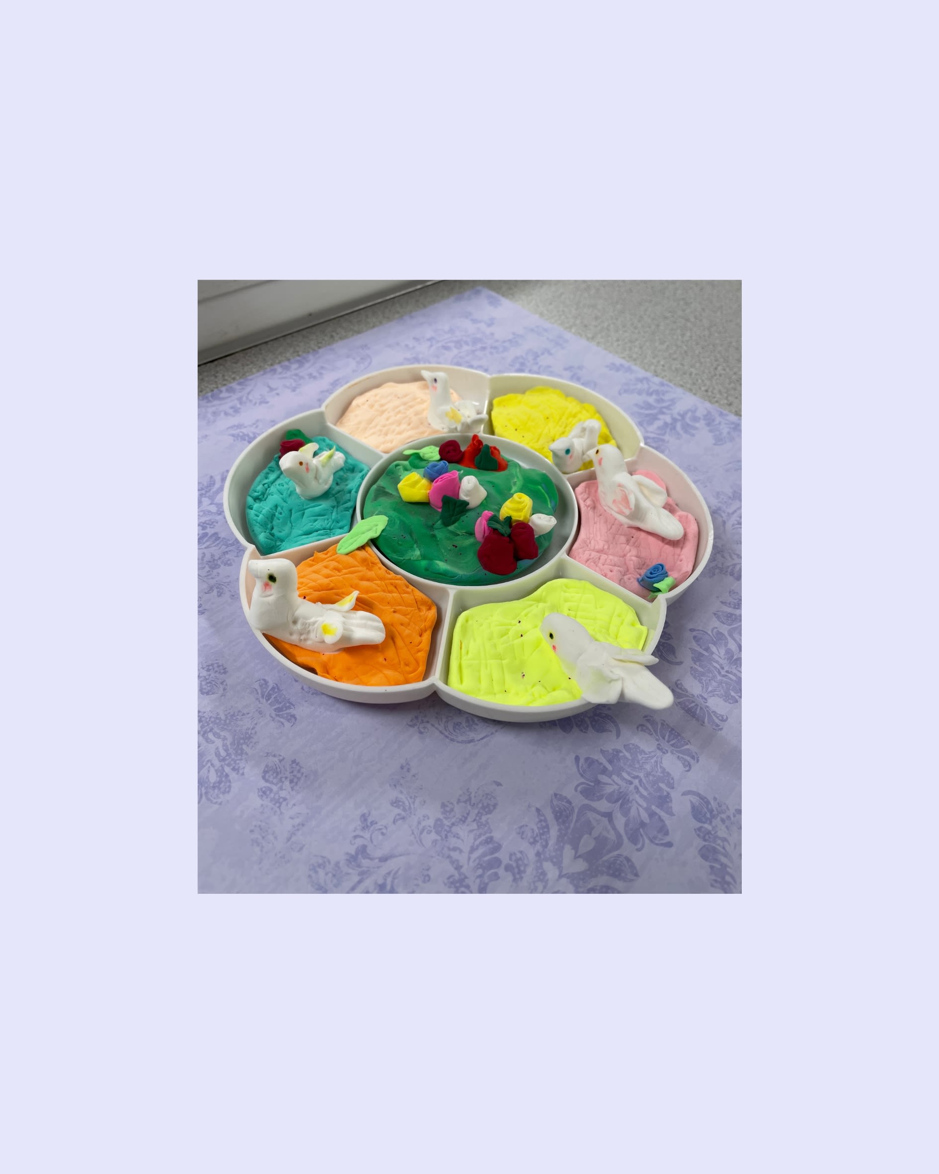 A clay figure created inside of a flower-shaped paint palette. Along the outer edge of the palette, there are white birds sitting on different colours of clay. In the middle of the palette is green clay with clay flowers on top.