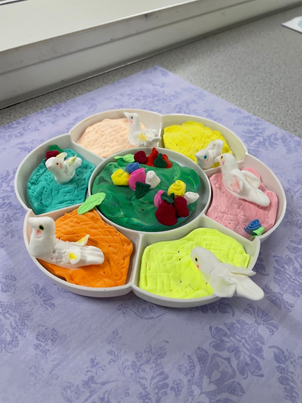 A clay figure created inside of a flower-shaped paint palette. Along the outer edge of the palette, there are white birds sitting on different colours of clay. In the middle of the palette is green clay with clay flowers on top.