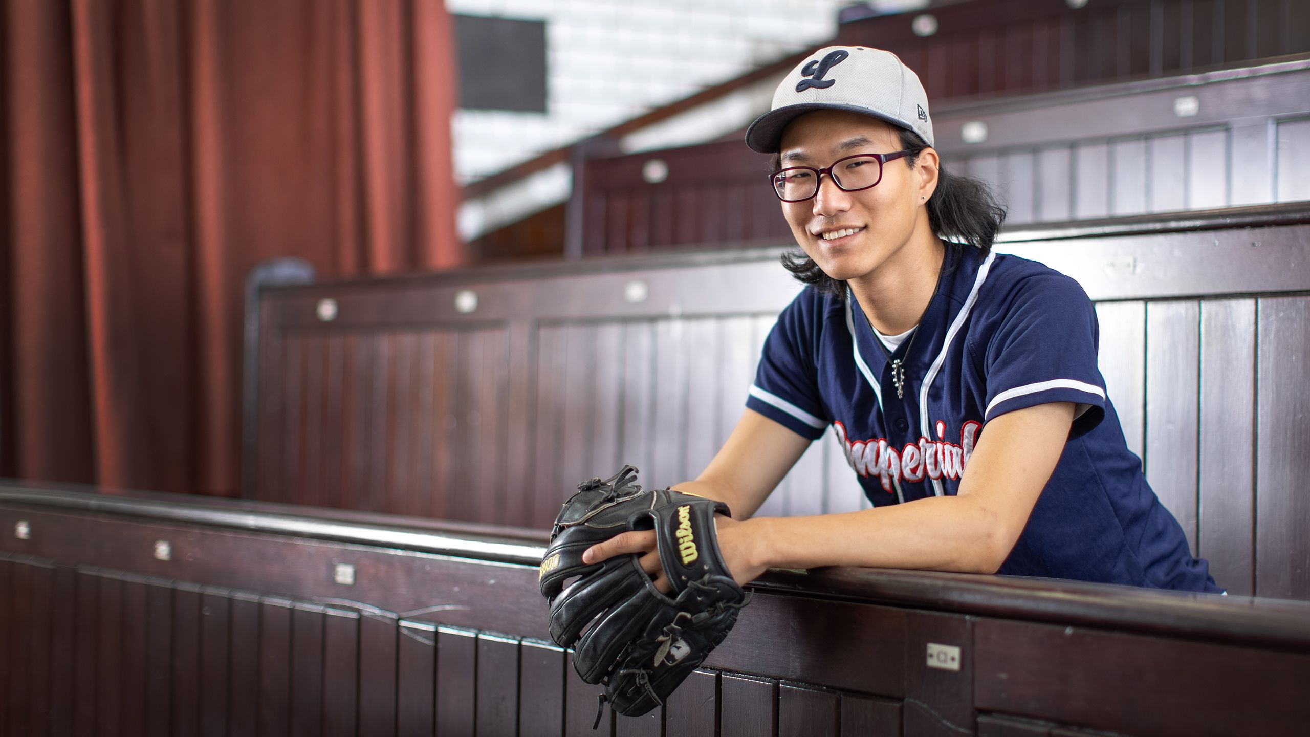 Ben Shin sits in lecture theatre wearing baseball outfit (hat and gloves) and smiles at the camera