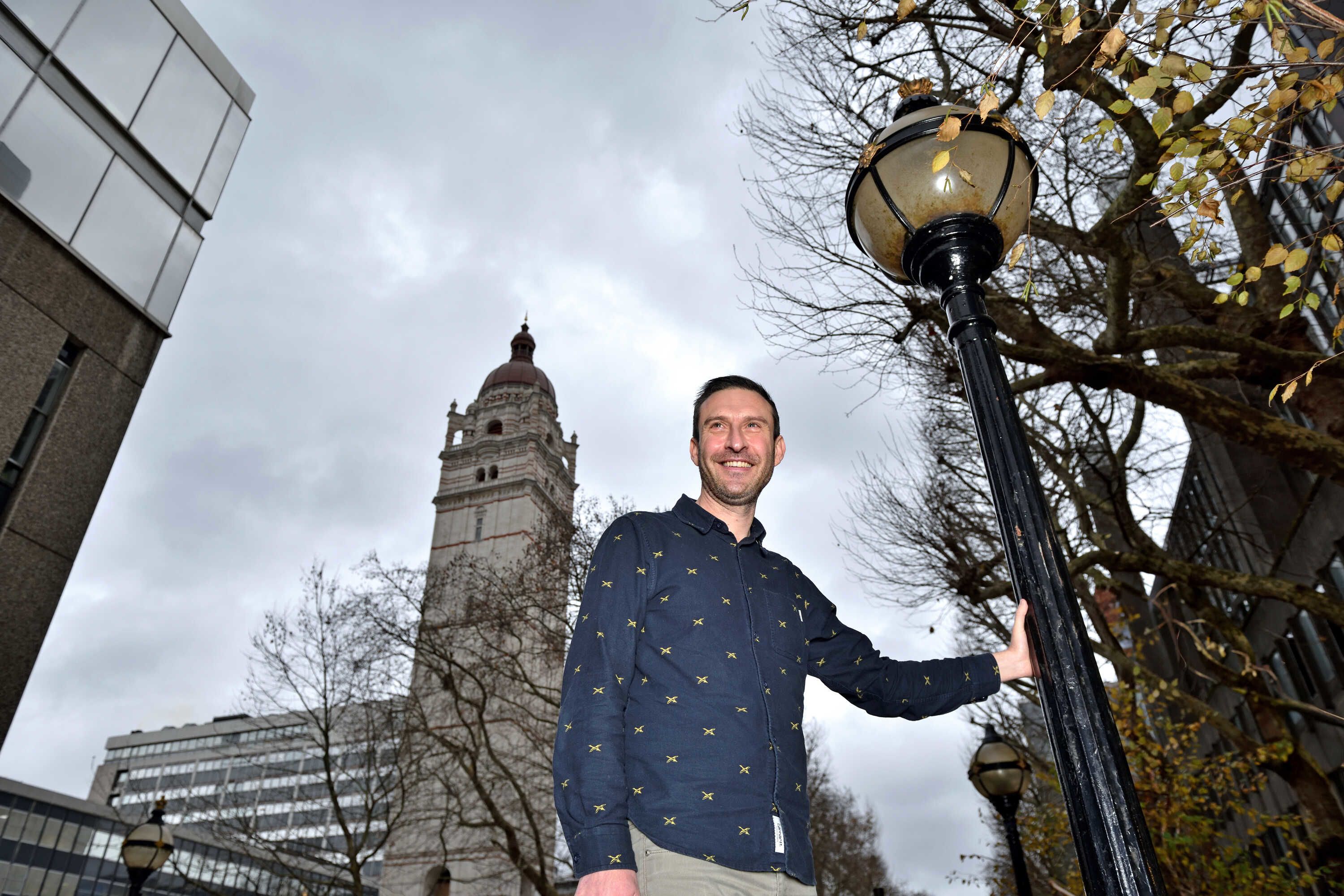 Photo from a low angle of Simon leaning on a lamppost in front of the Queens Tower.