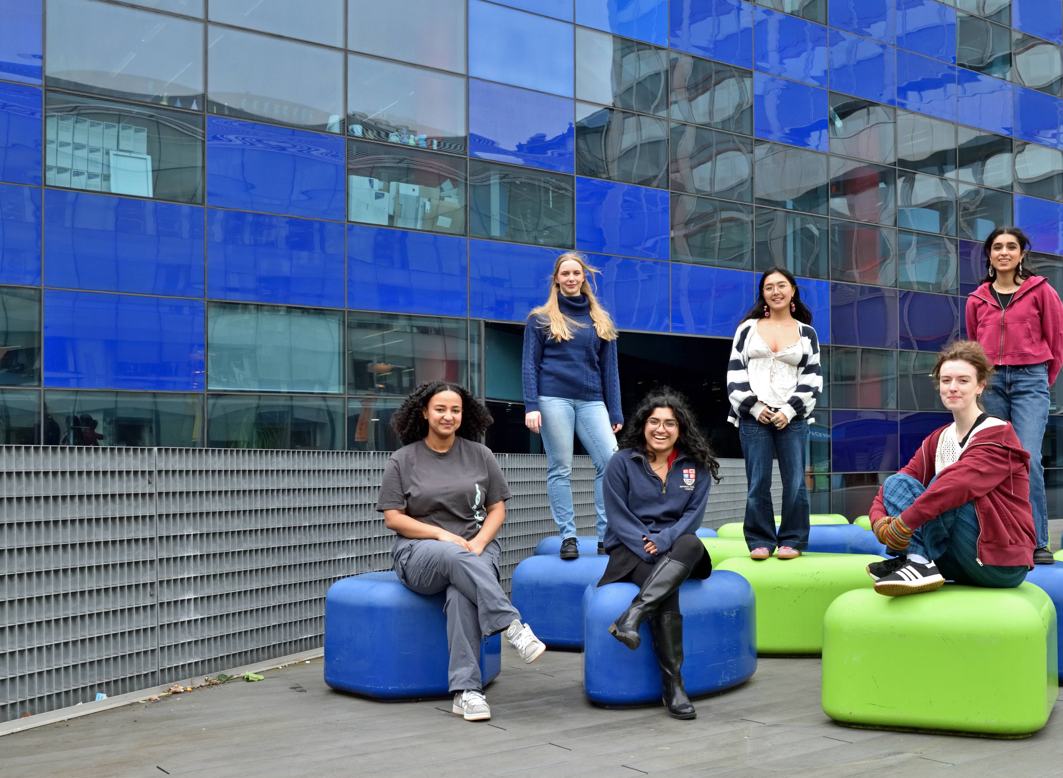 Six members of the society posing for a photo, three of them sitting on green and blue stools in the foreground, and three of them standing on the stools in the background.