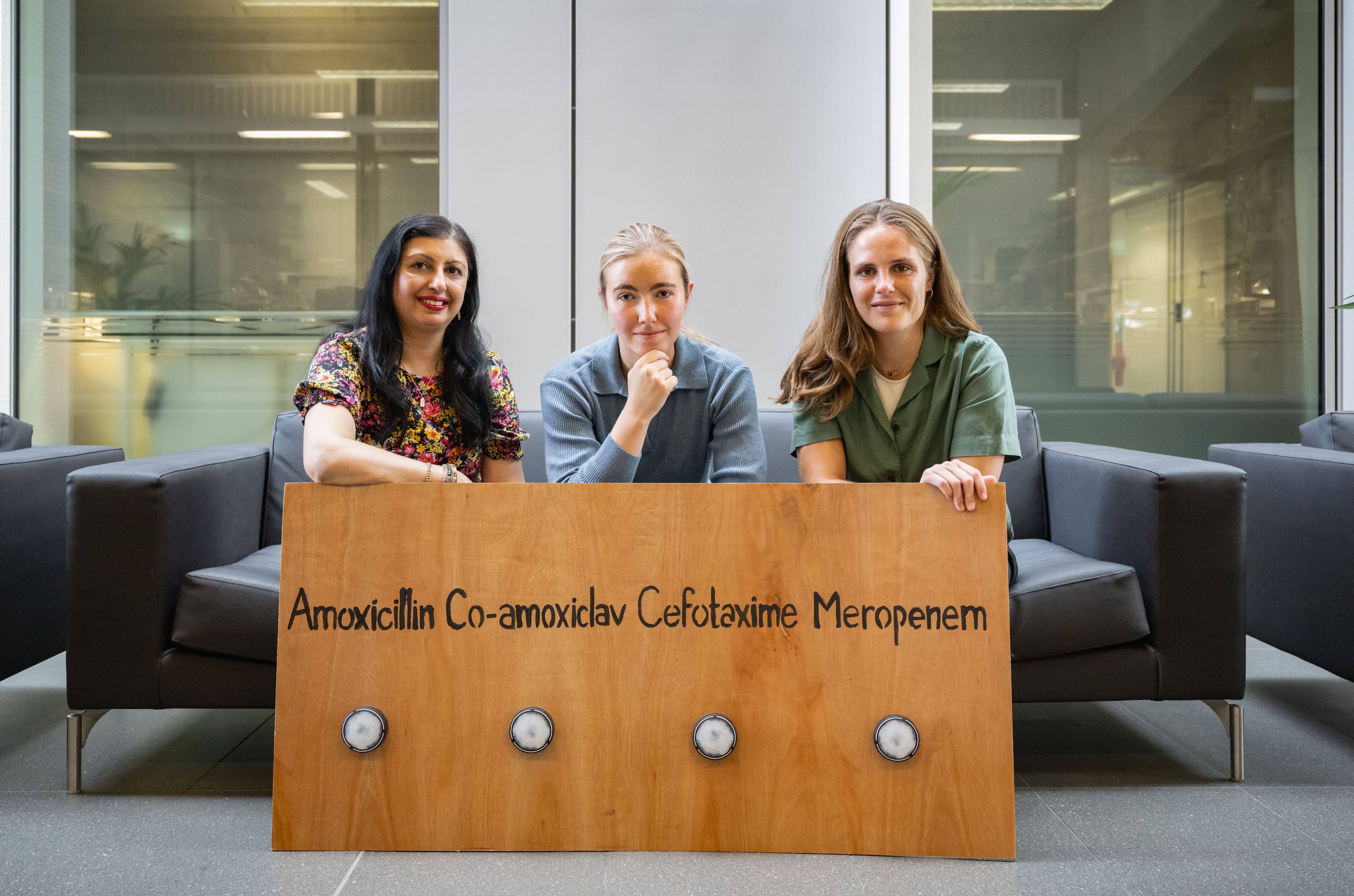 Rakhee, Jenny and Ellen sitting behind a sign with the names of three types of bacteria, which the team used in one of their engagement activities